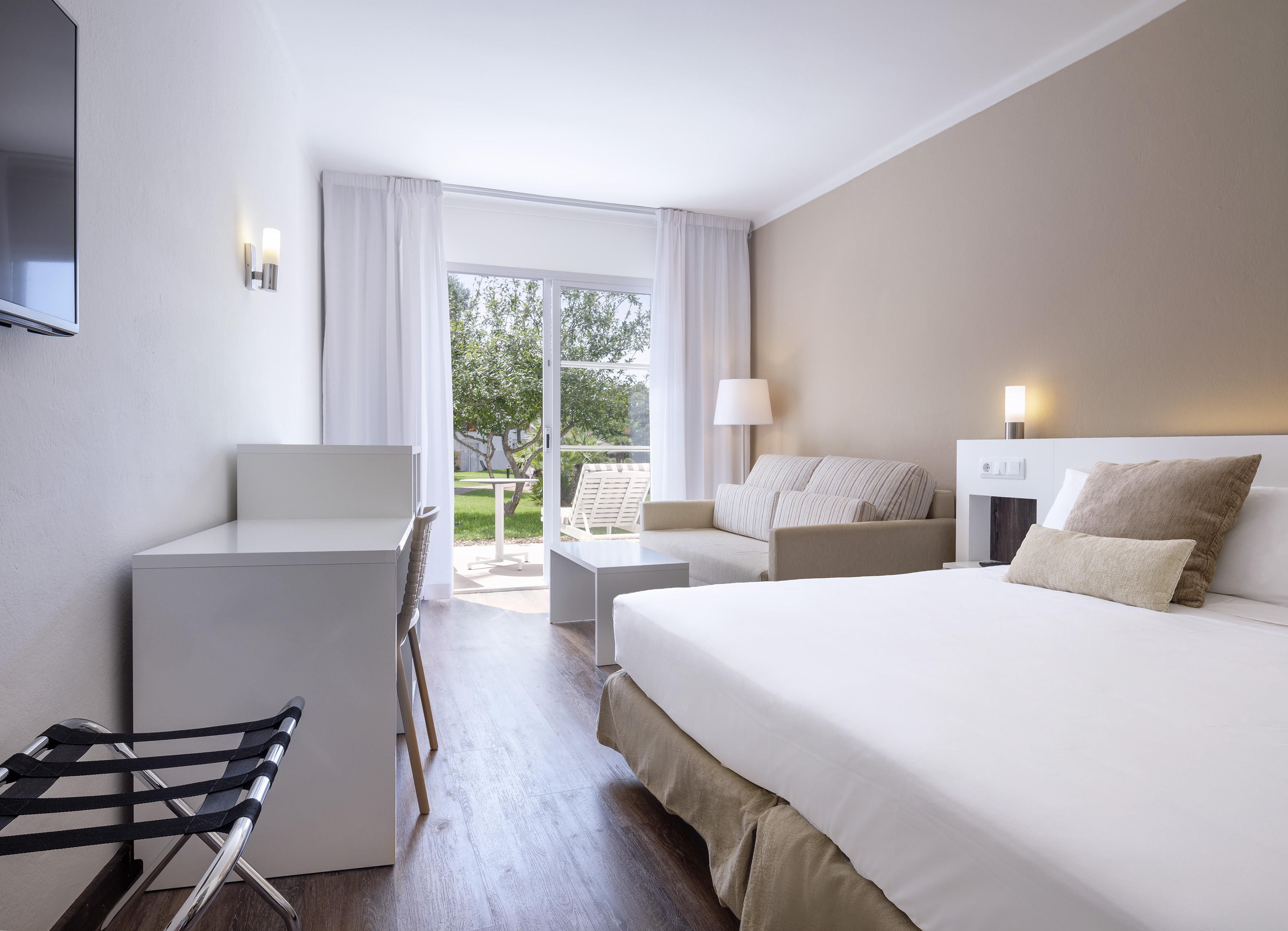 Modern hotel room with bed, sofa, desk, and garden view patio. - XTRA SOL Suite - SOL Falcó Menorca, Menorca