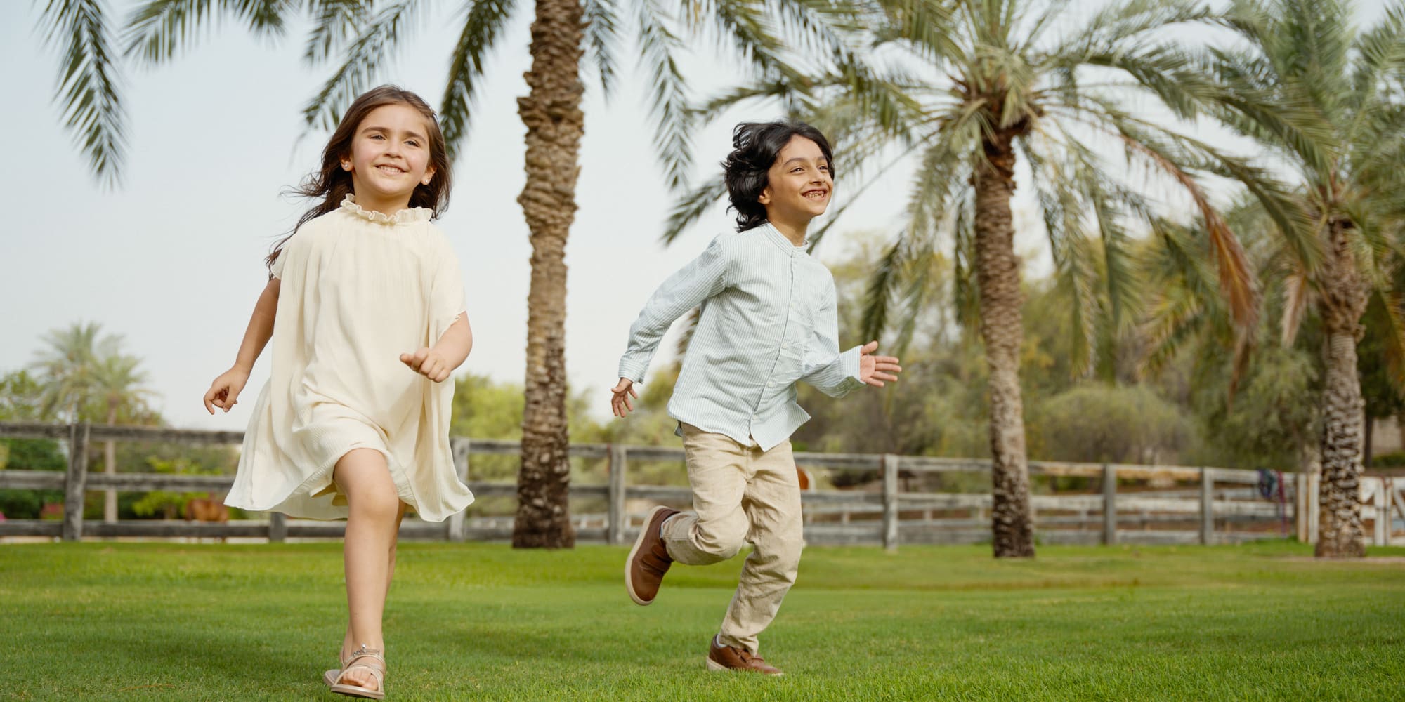 Two smiling kids playing on lush green grass with tall palm trees.