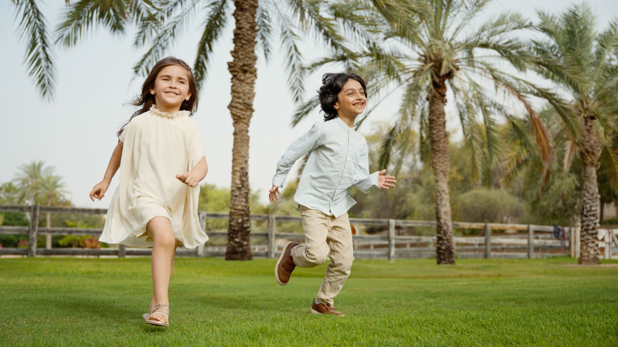 Two smiling kids playing on lush green grass with tall palm trees.
