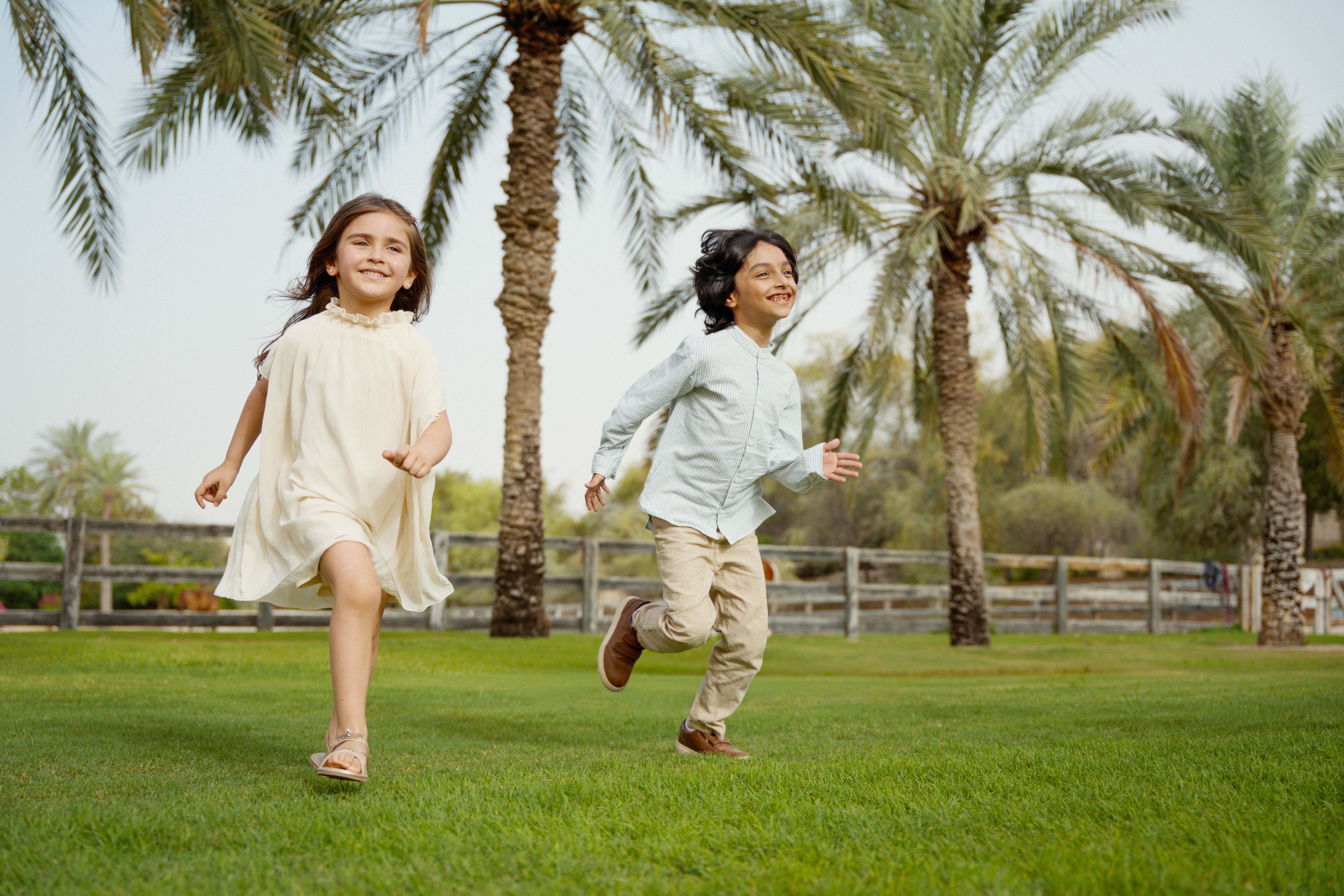 Two smiling kids playing on lush green grass with tall palm trees.
