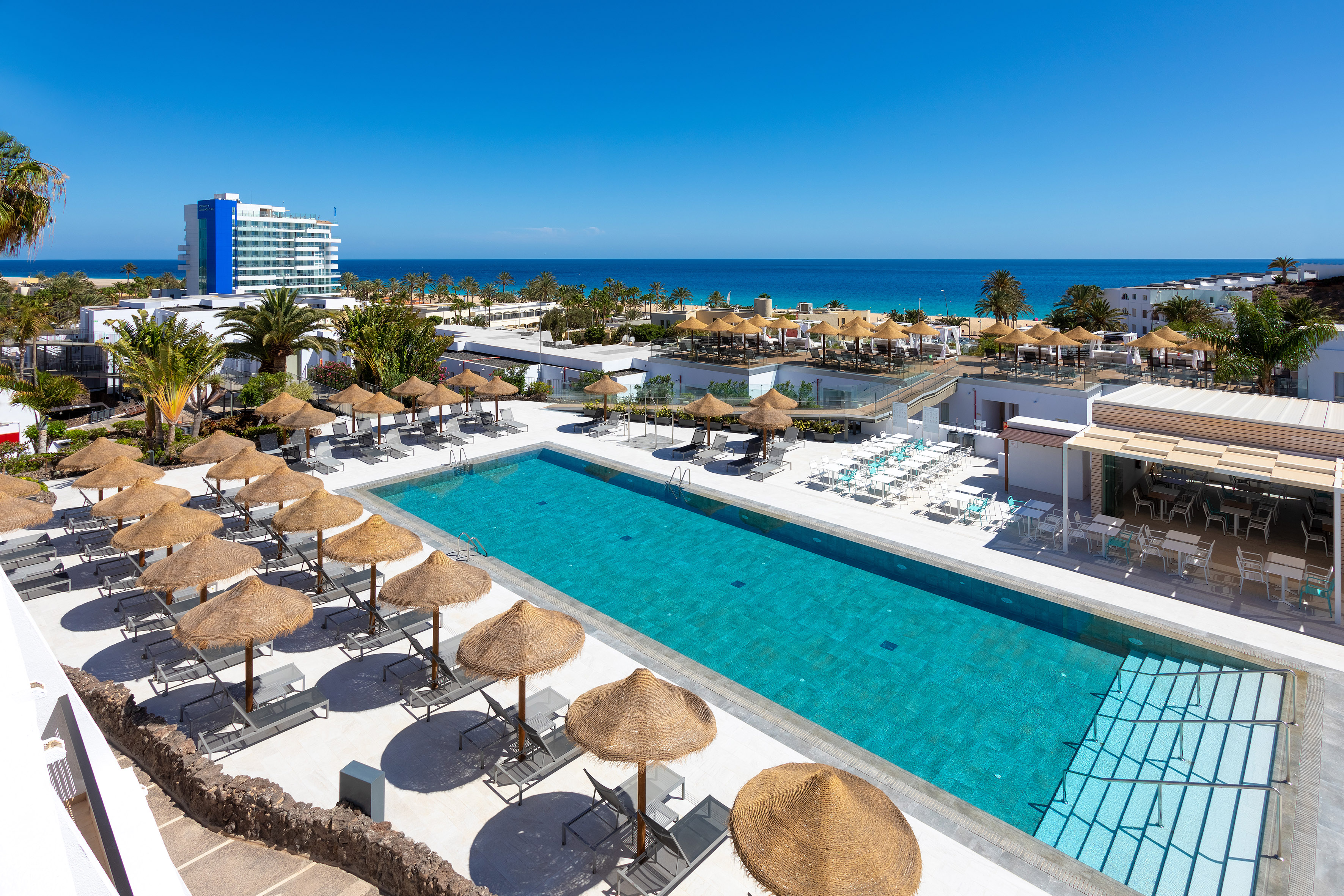 Resort swimming pool with sun loungers, straw umbrellas, ocean, and beach.