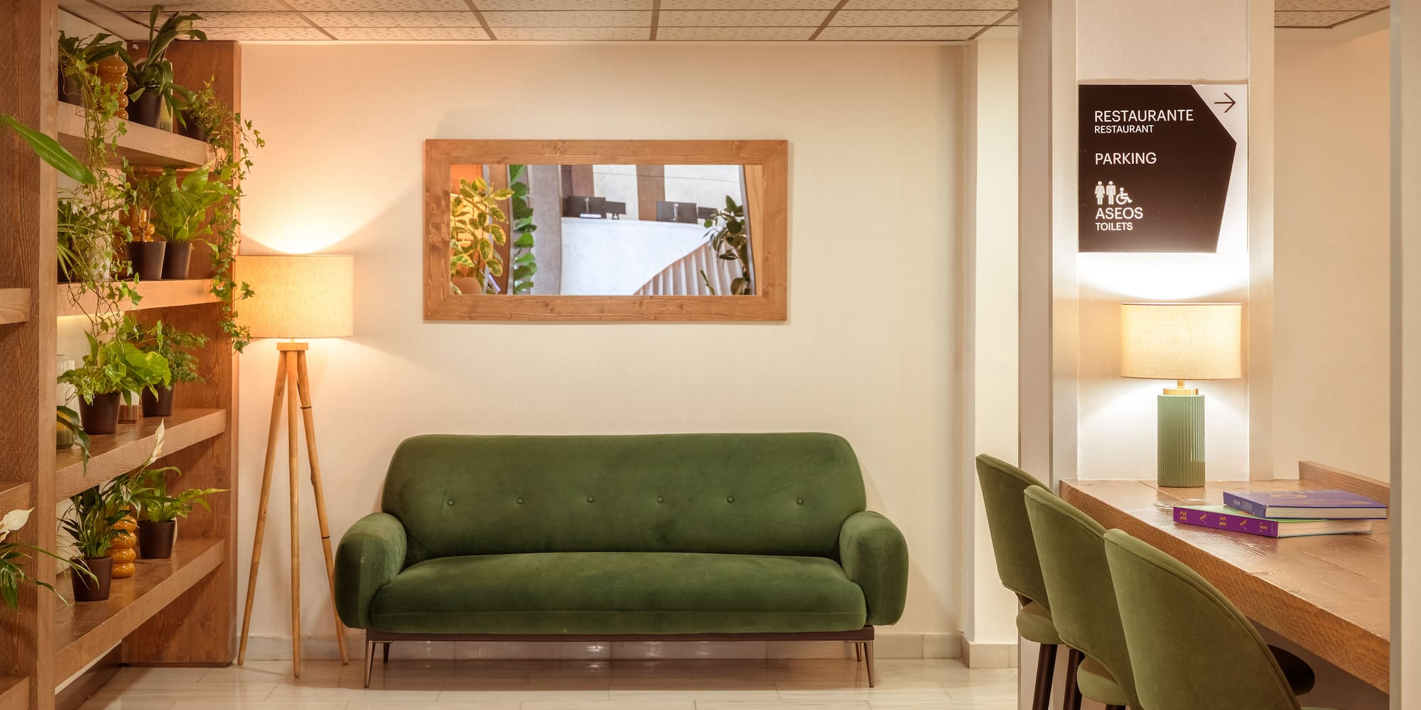Cozy hotel lounge with green sofa, plants, mirror, and amenities sign.