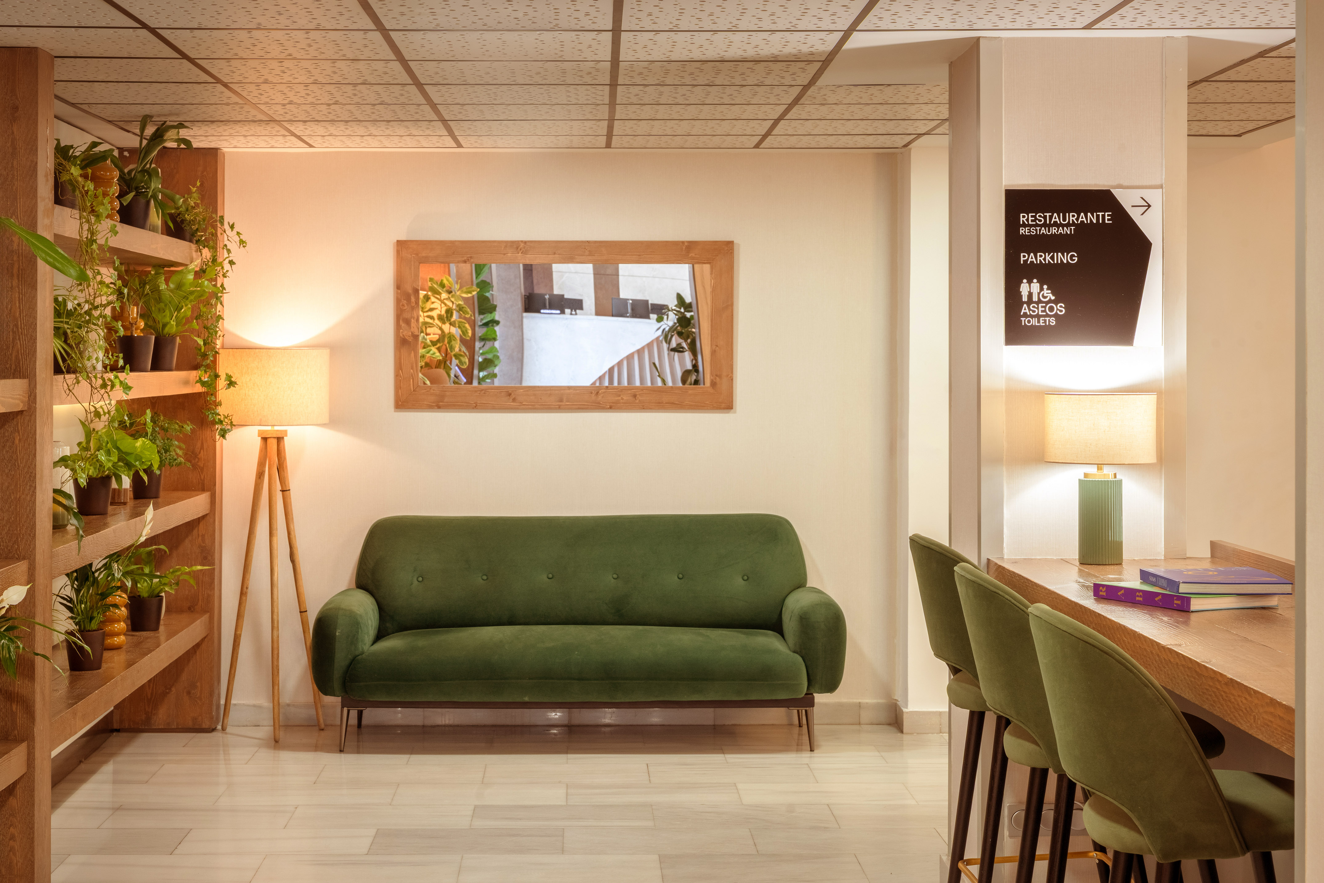 Cozy hotel lounge with green sofa, plants, mirror, and amenities sign.