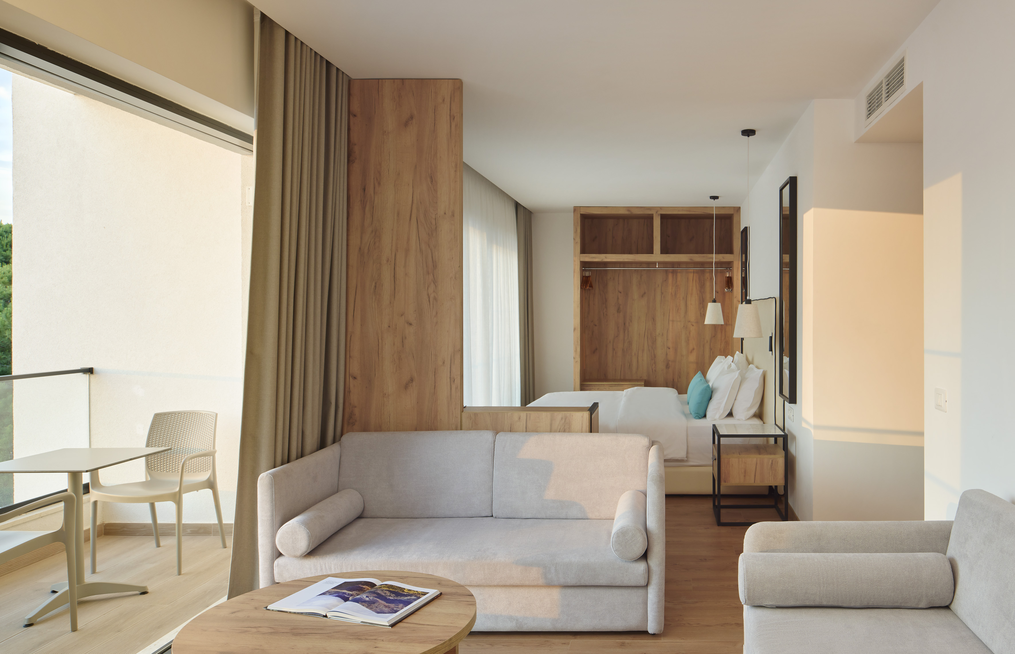 Modern hotel room suite with living area, bed, wood accents, and sunlit balcony.