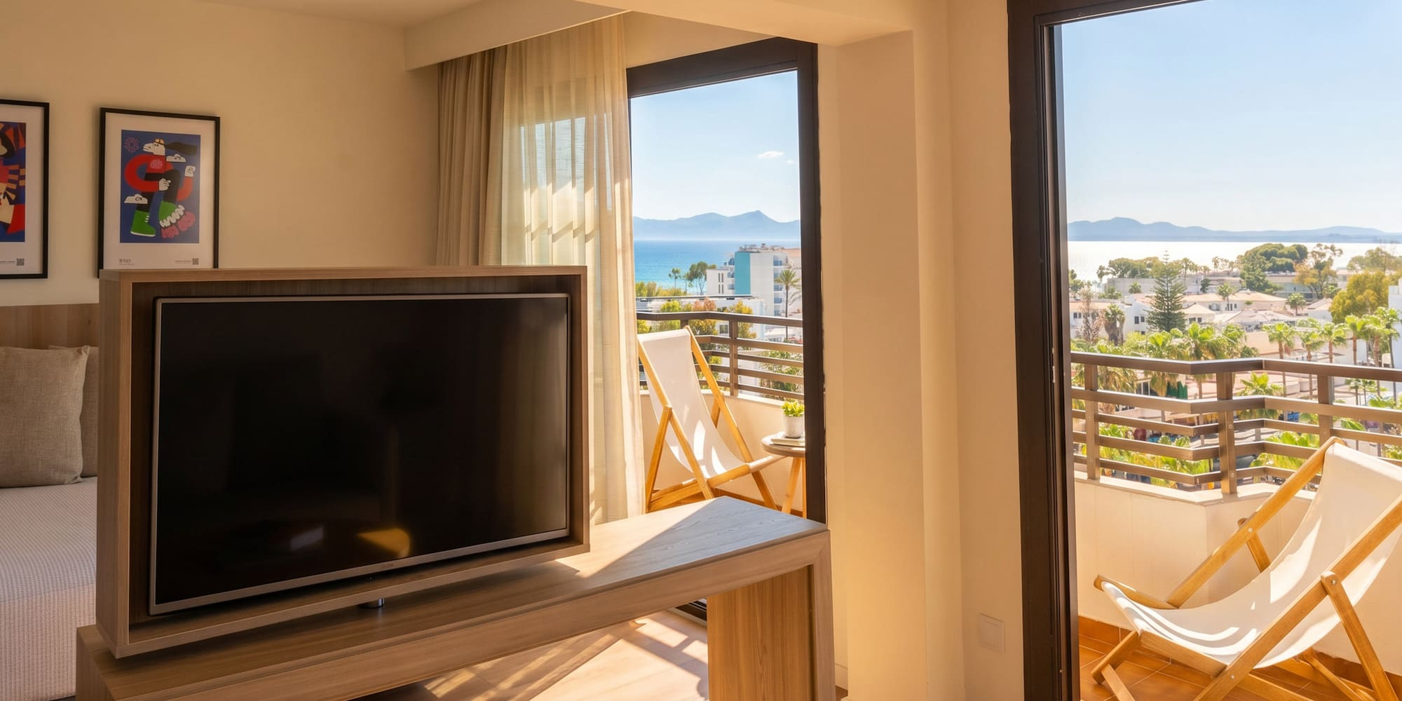 Sunny hotel room with TV, balcony deck chairs, and panoramic sea view.