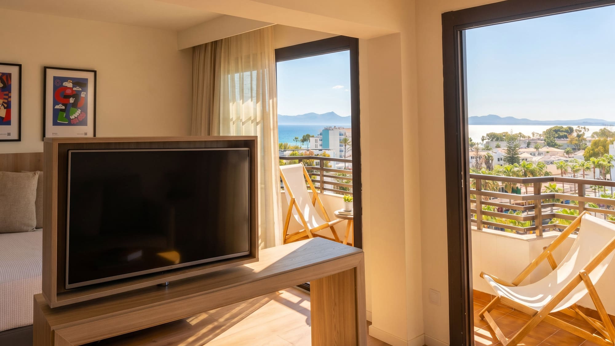 Sunny hotel room with TV, balcony deck chairs, and panoramic sea view.