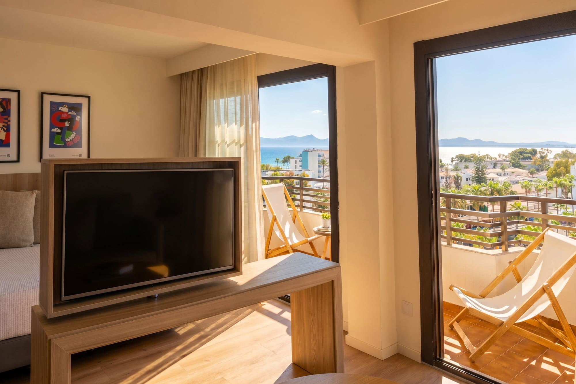 Sunny hotel room with TV, balcony deck chairs, and panoramic sea view.