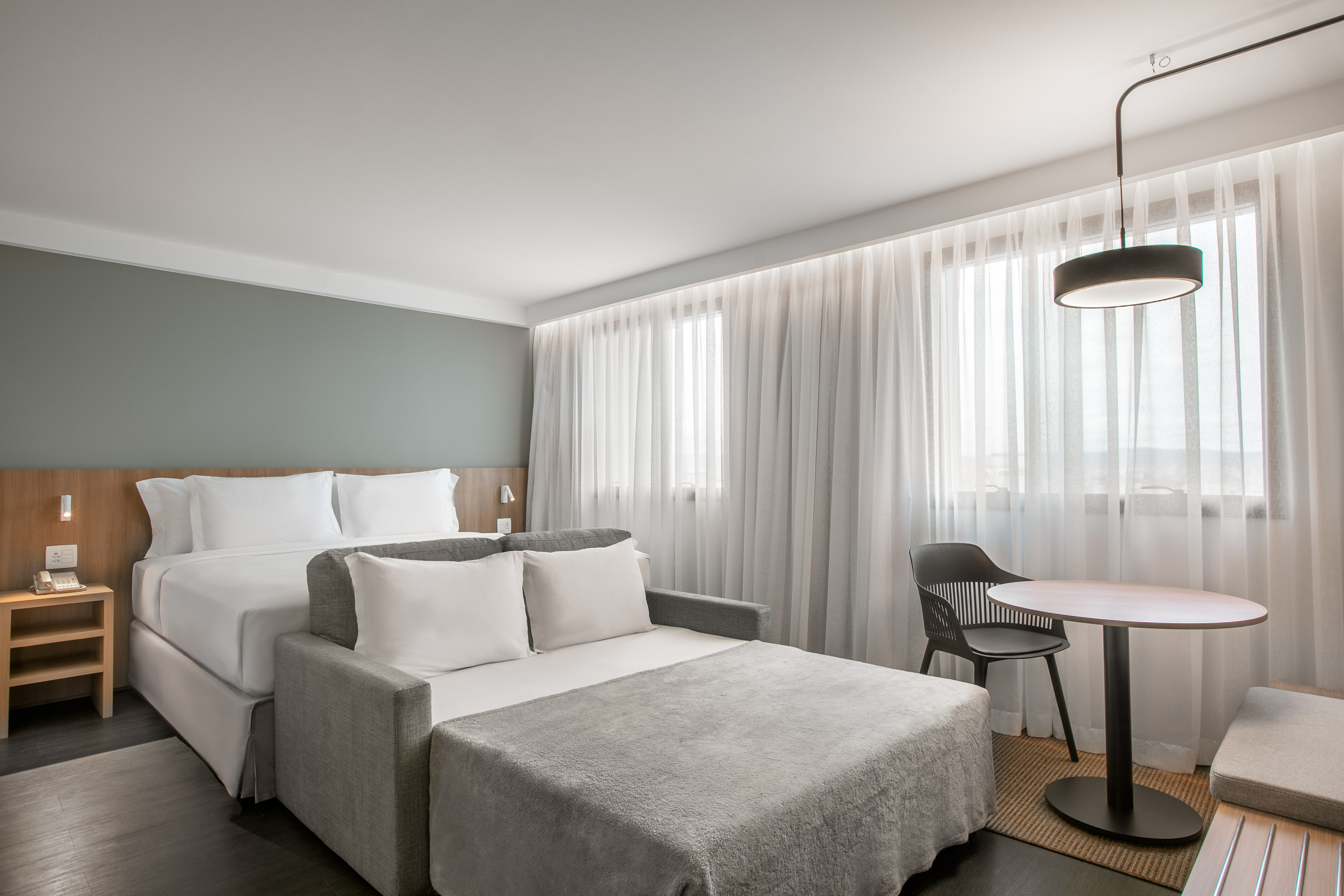 Modern hotel room with bed, sofa bed, nightstand, and table by window.