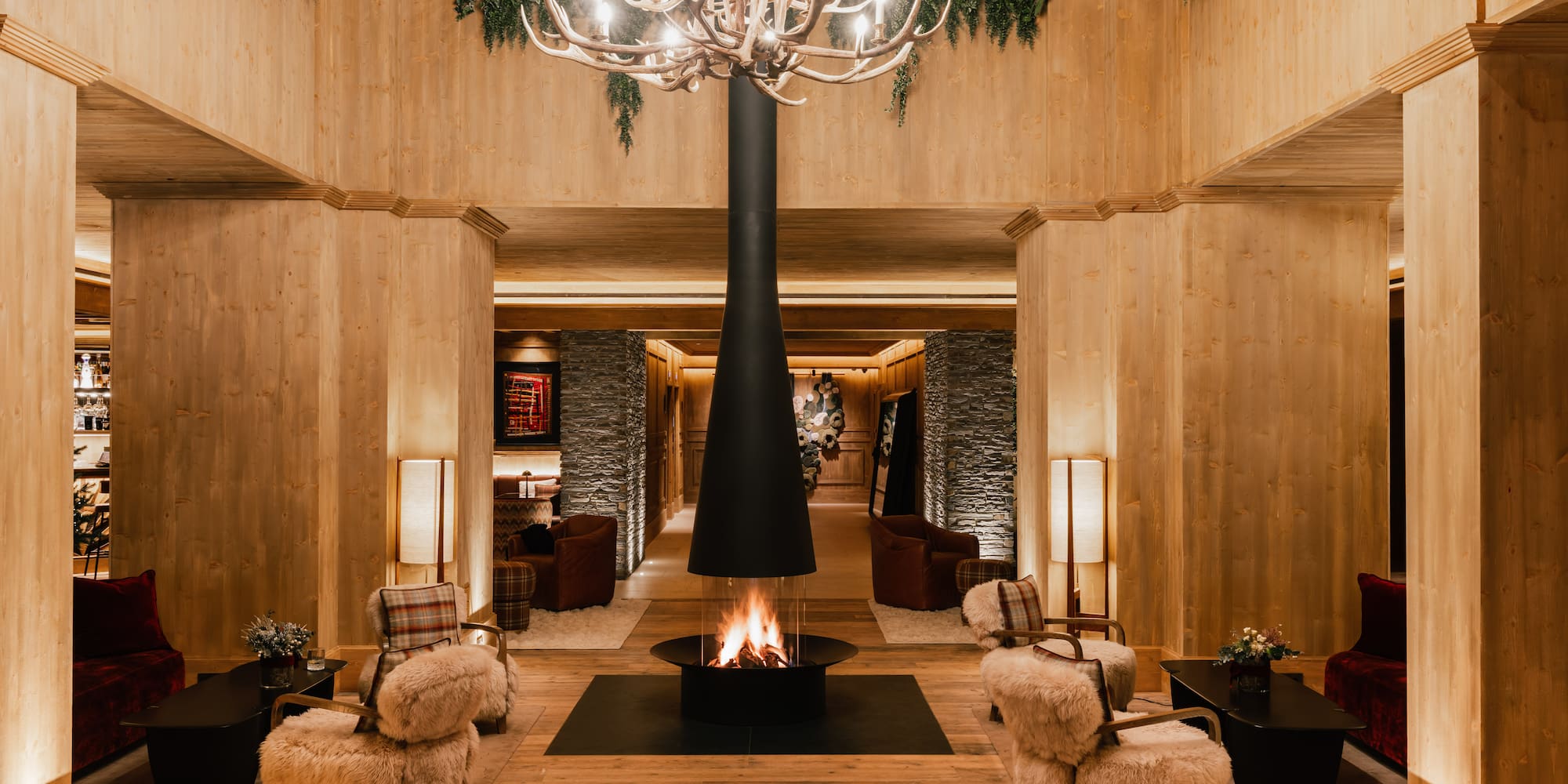 Lobby de hotel alpino con chimenea central, madera y candelabro de astas.