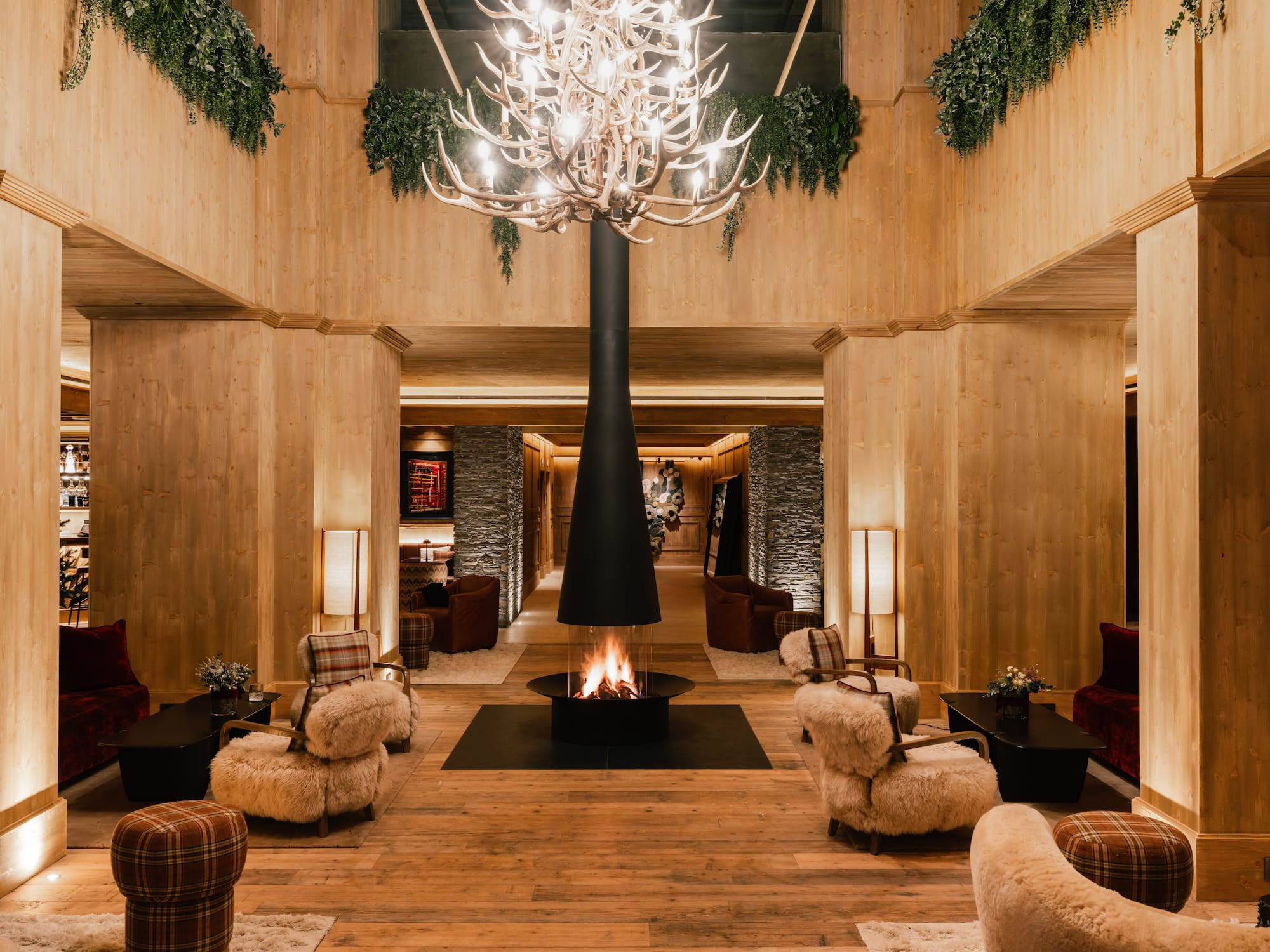 Lobby de hotel alpino con chimenea central, madera y candelabro de astas.