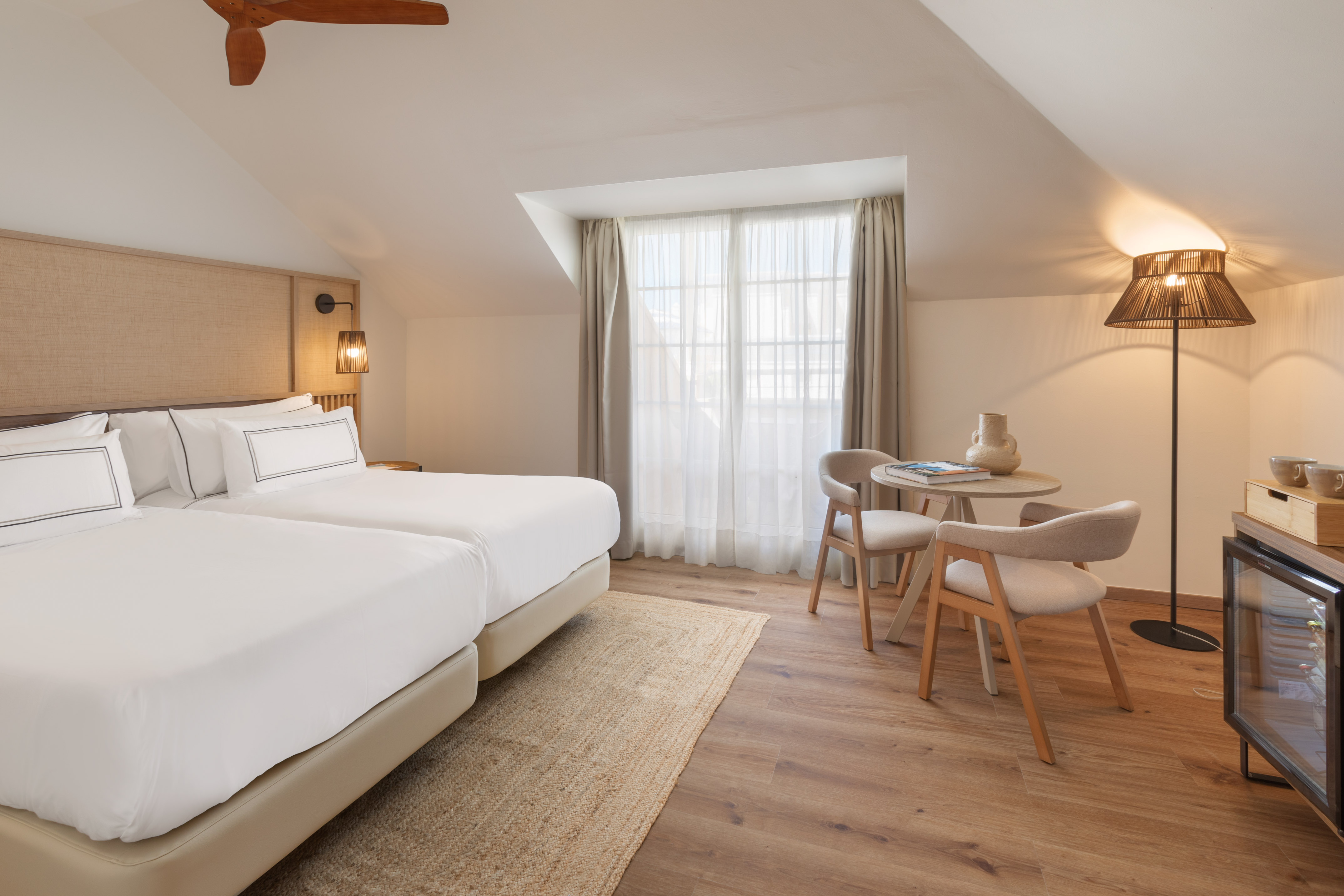 Bright attic hotel room with two twin beds, table, and wooden ceiling fan.
