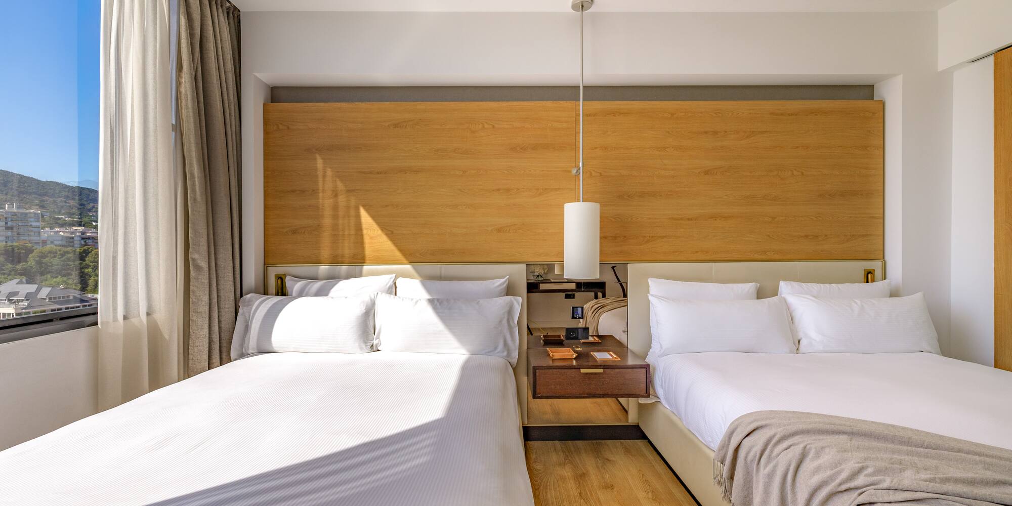 Modern hotel room with two beds, wooden headboard, and scenic mountain view.