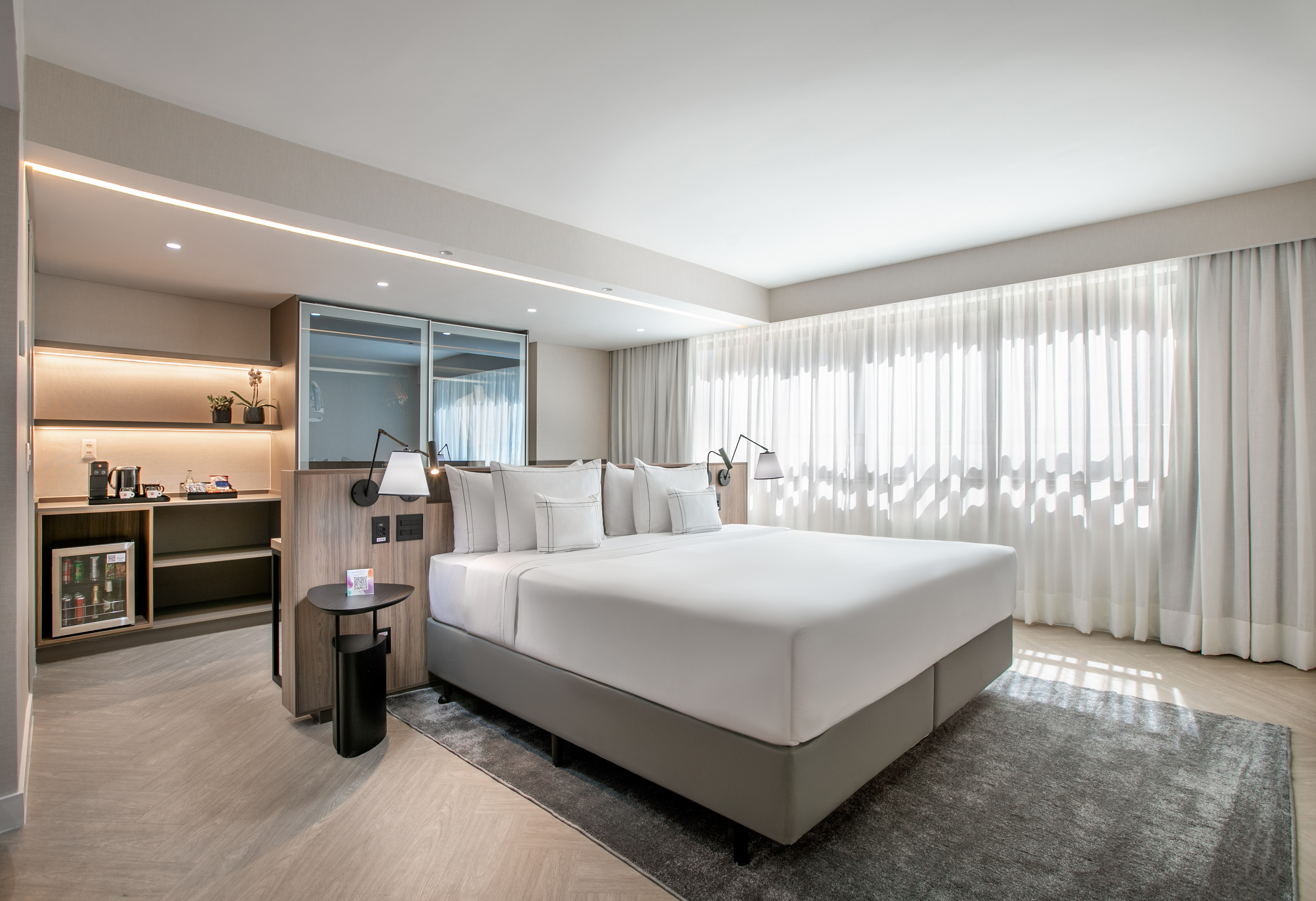 Modern hotel room with king bed, minibar, and sheer curtains.