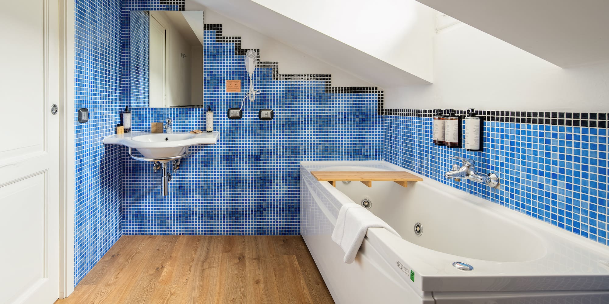 Modern attic bathroom with blue mosaic tiles, white bathtub, sink, and wood floor.