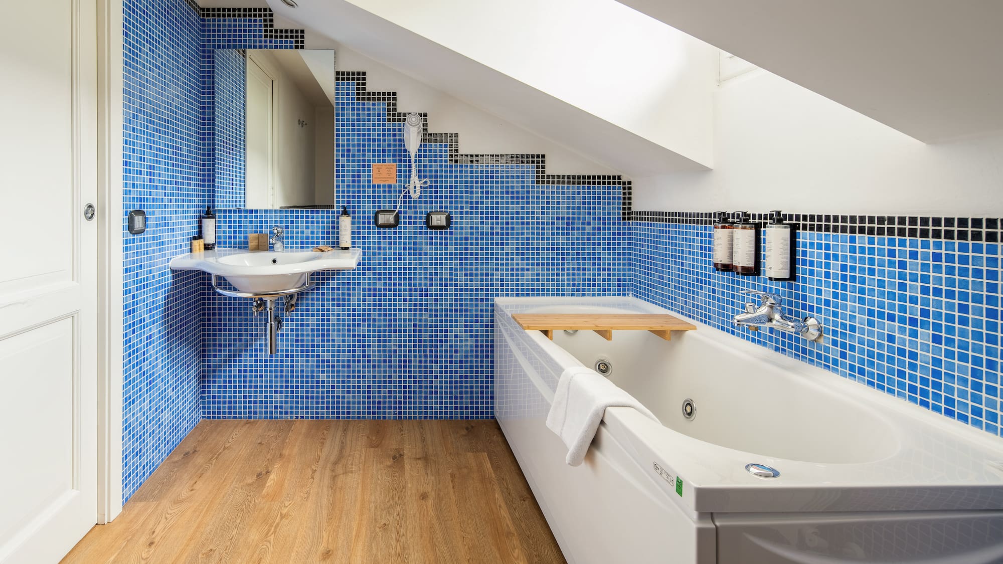 Modern attic bathroom with blue mosaic tiles, white bathtub, sink, and wood floor.