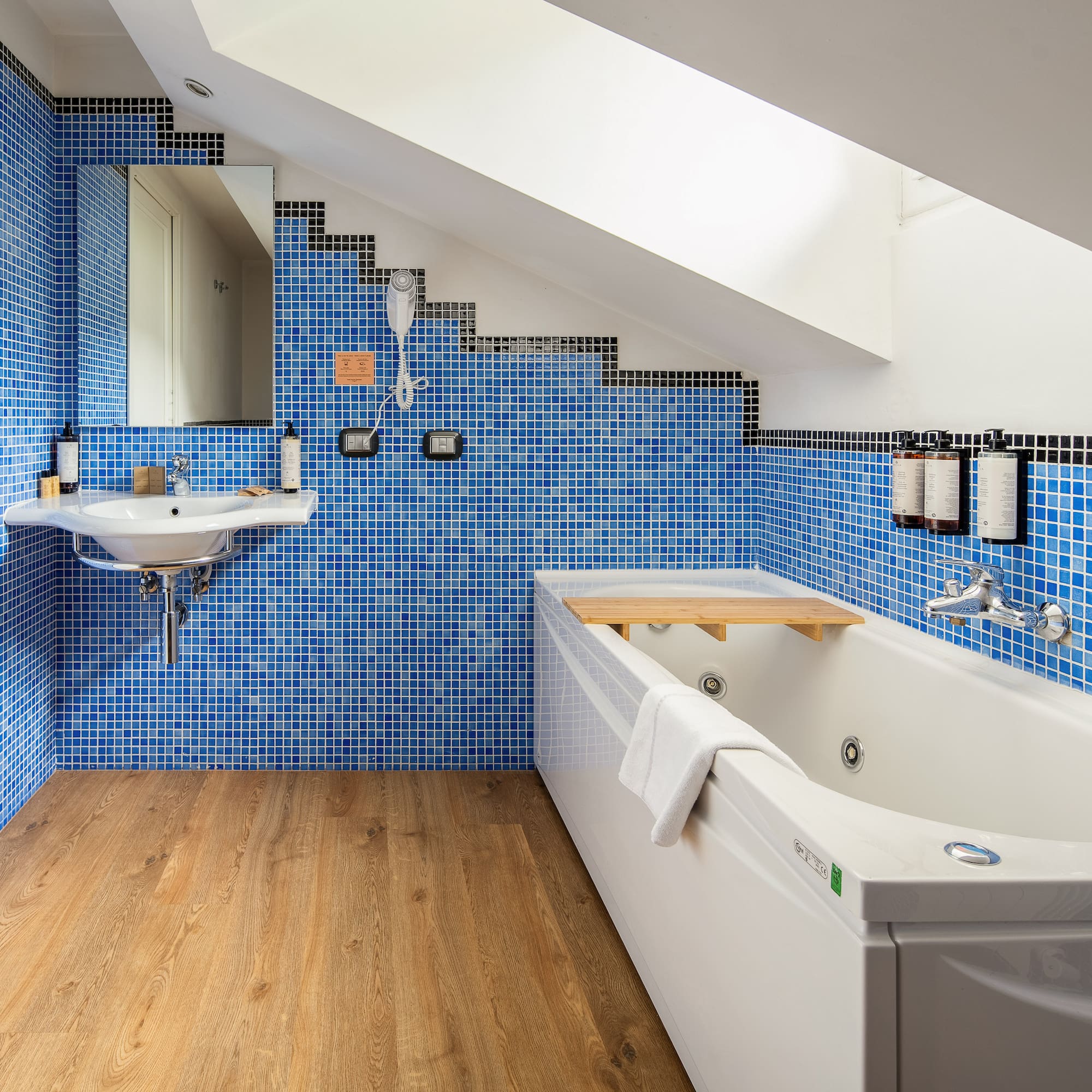 Modern attic bathroom with blue mosaic tiles, white bathtub, sink, and wood floor.