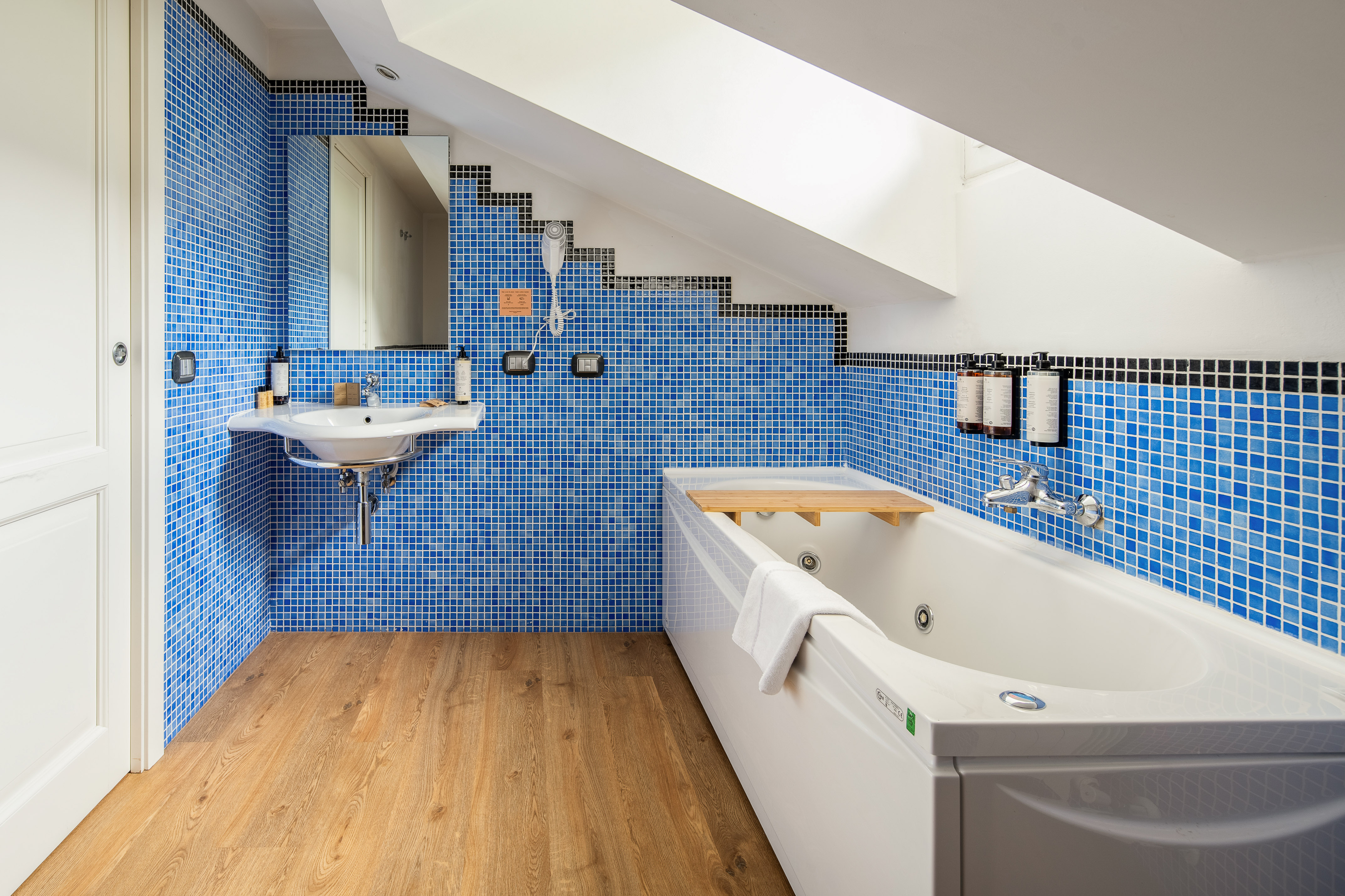 Modern attic bathroom with blue mosaic tiles, white bathtub, sink, and wood floor.