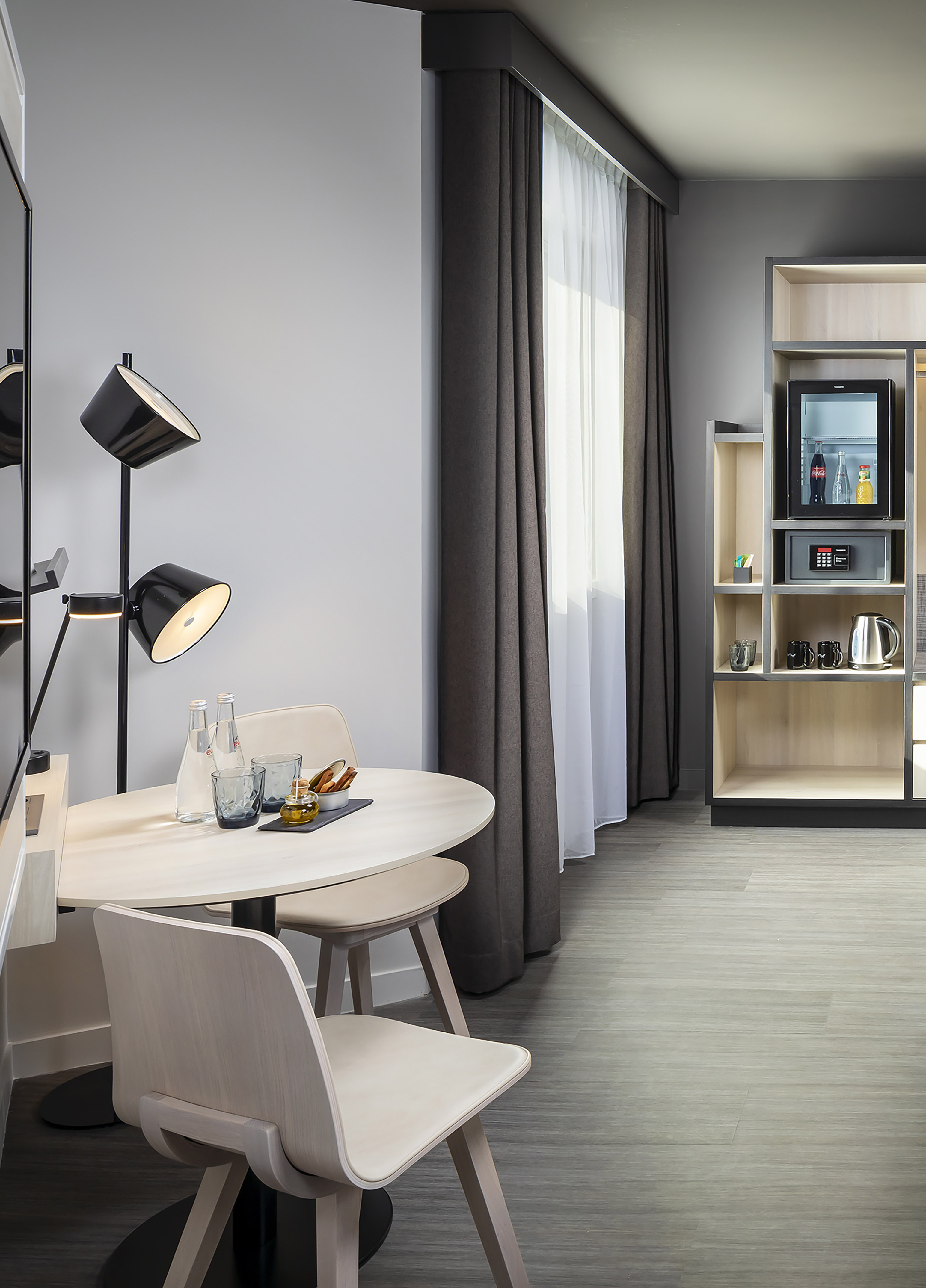 Modern hotel suite dining area with light table, chairs, black lamp, minibar, safe, and window.