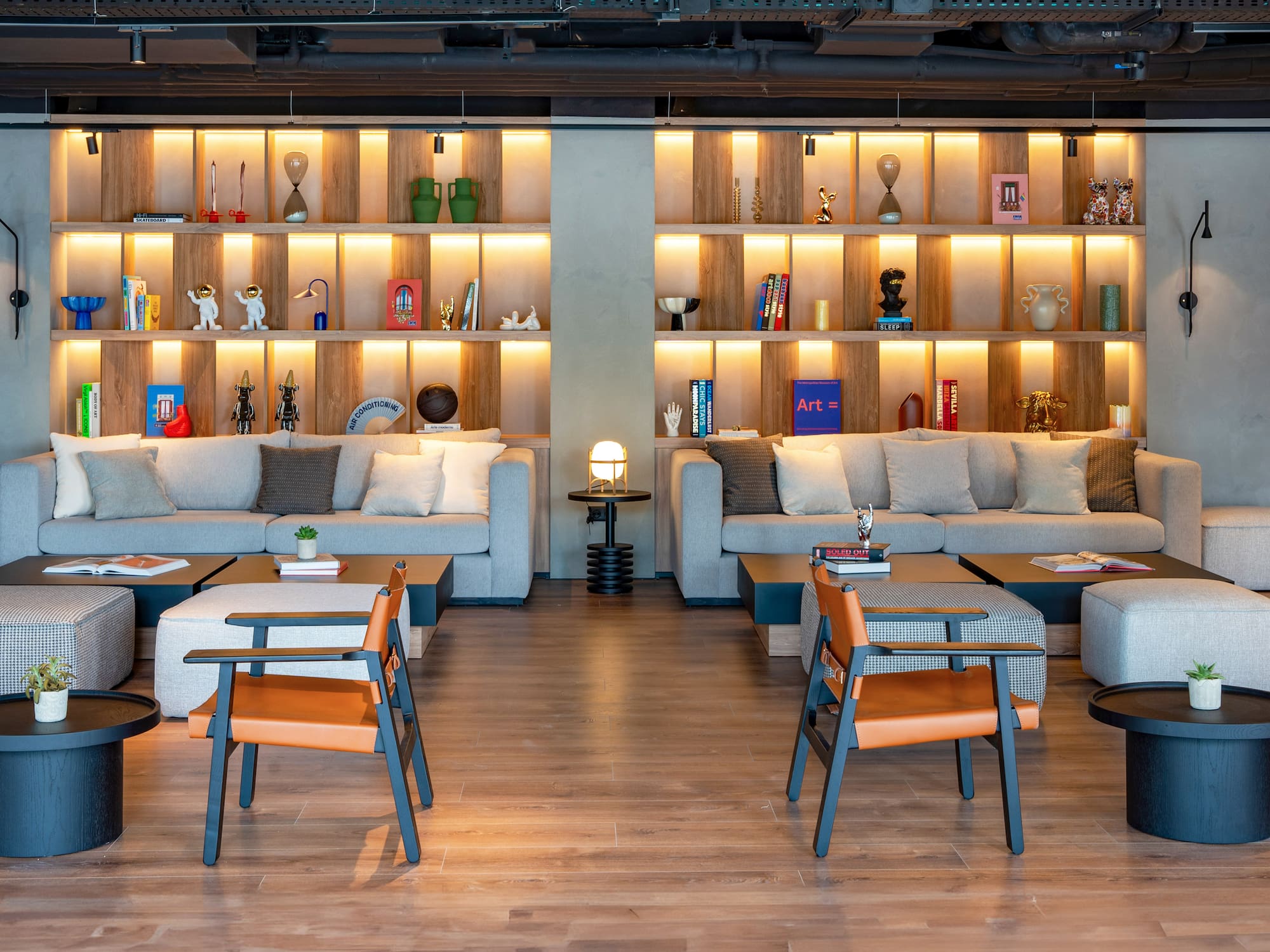 Modern hotel lounge with grey sofas, orange chairs, and backlit decorative shelves.