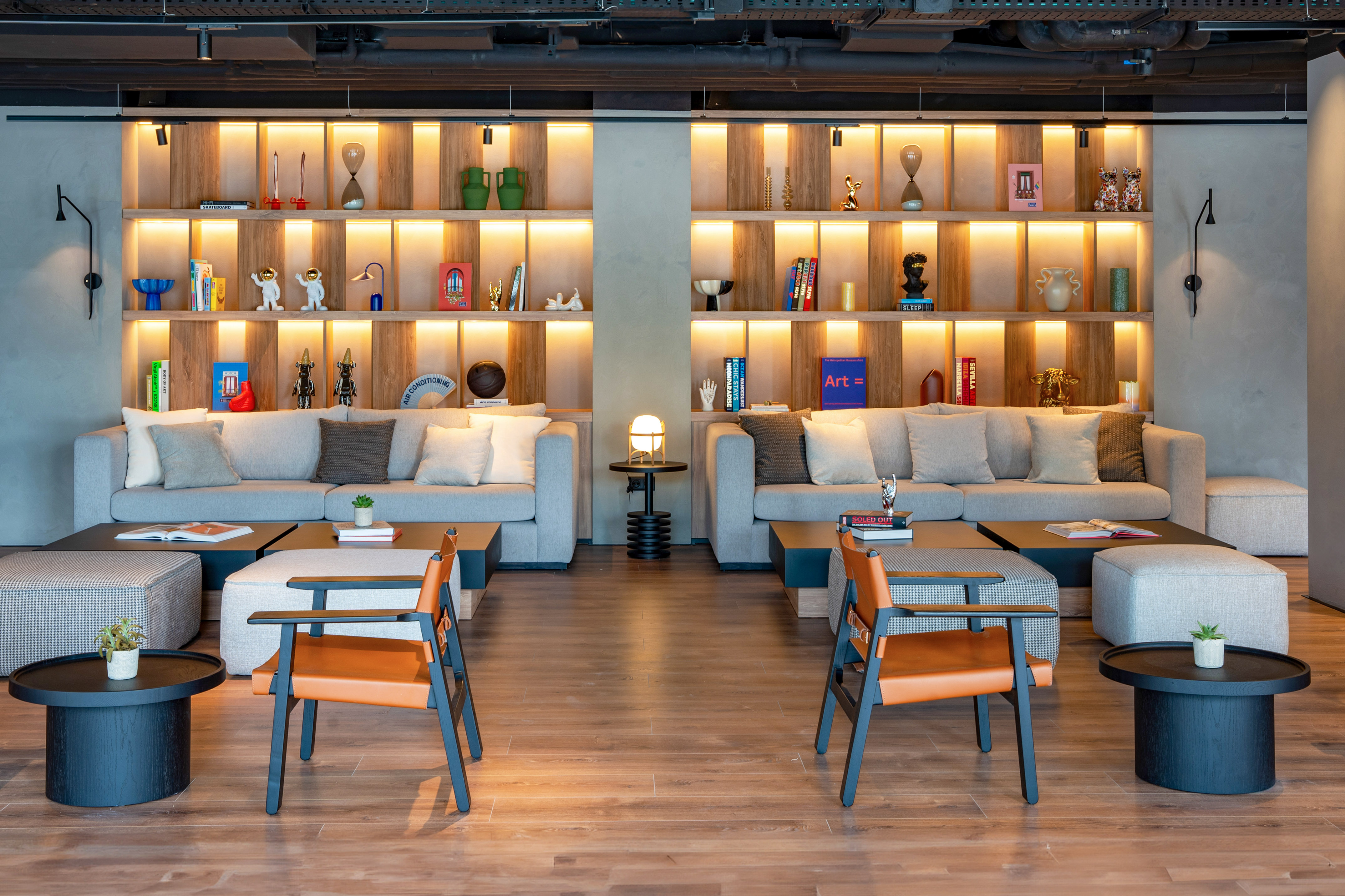 Modern hotel lounge with grey sofas, orange chairs, and backlit decorative shelves.