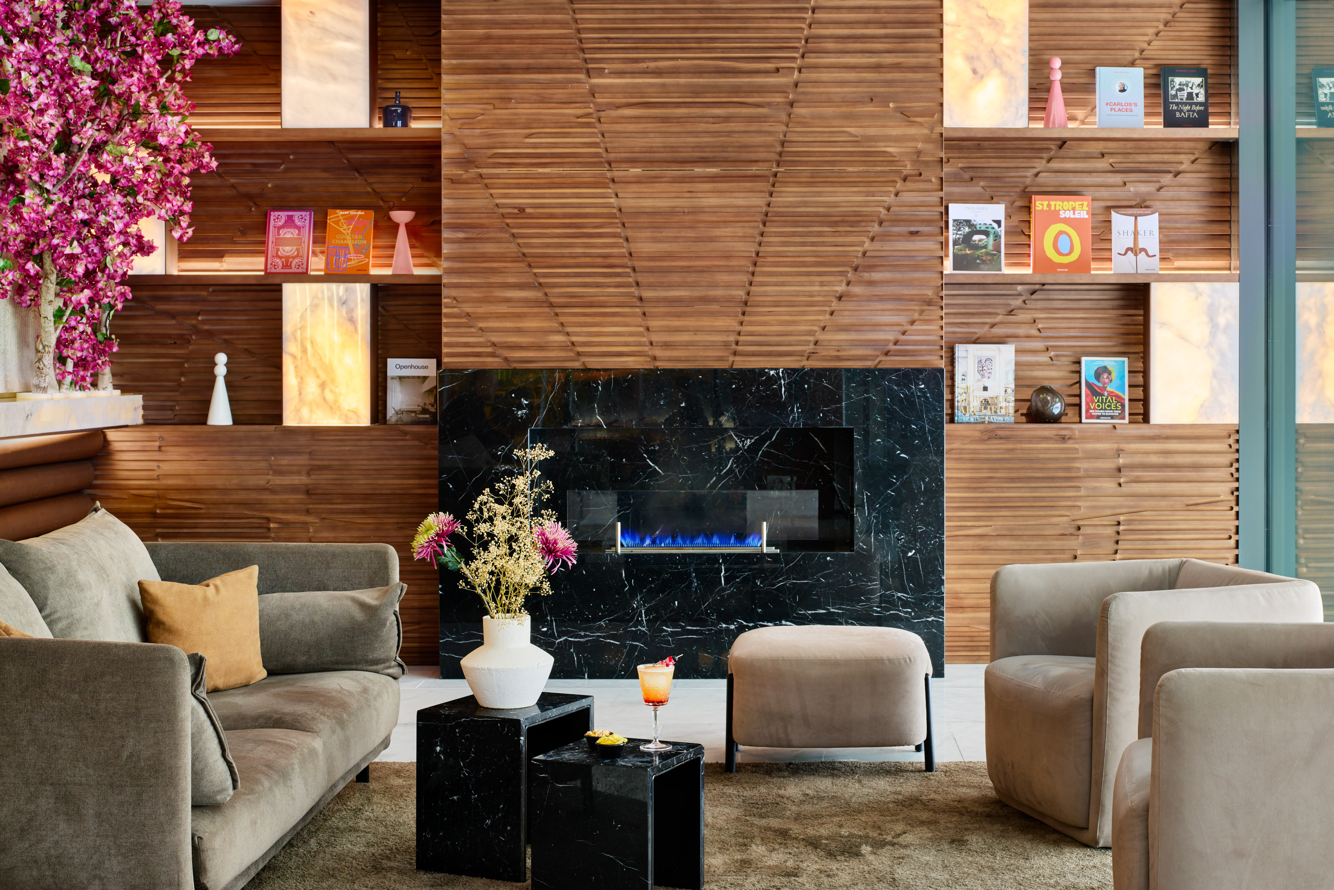 Modern lounge, black marble fireplace, wood shelves, pink flowers, cozy seating.