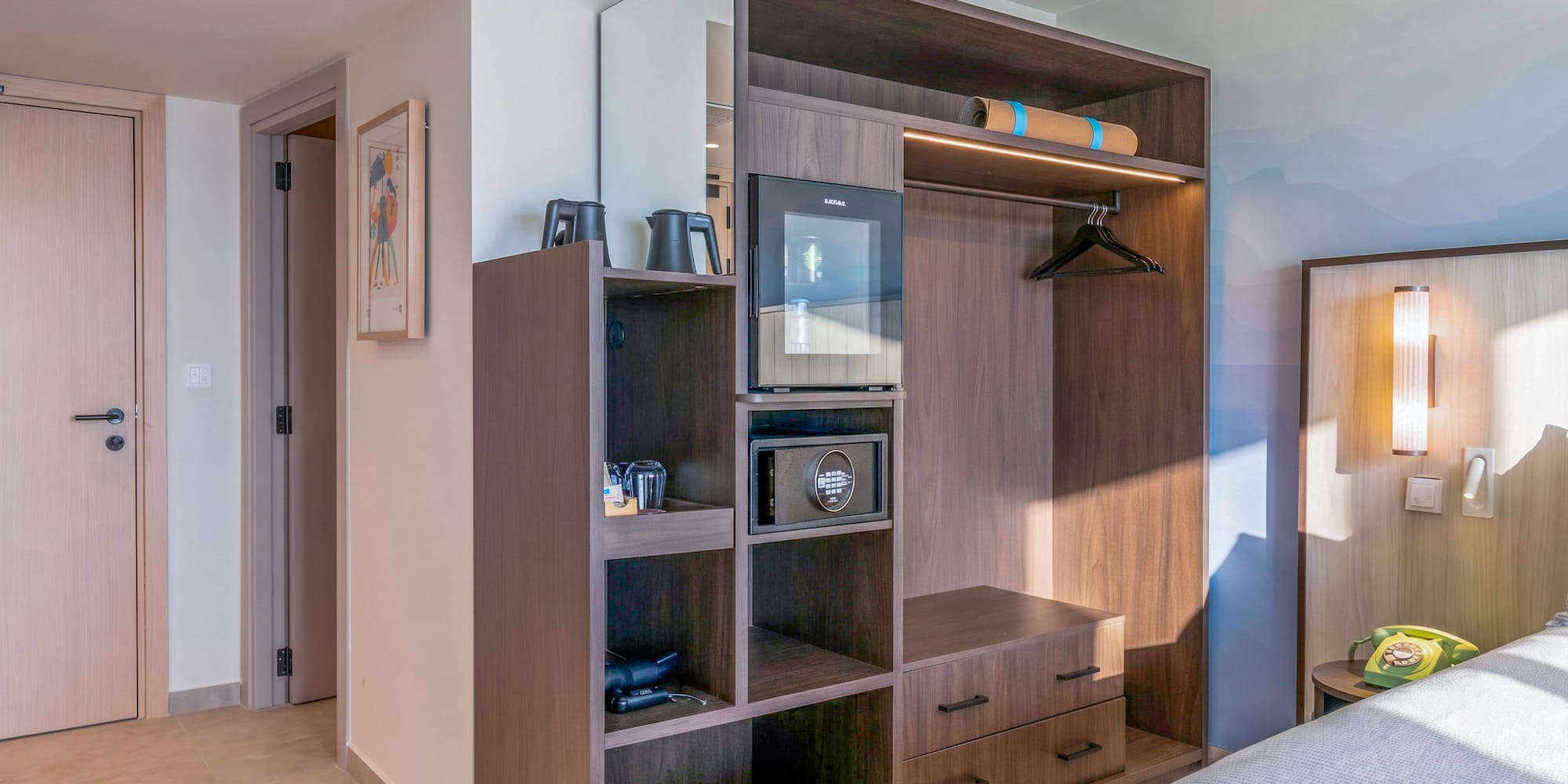 Modern hotel room with built-in dark wood wardrobe, mini-fridge, and safe.
