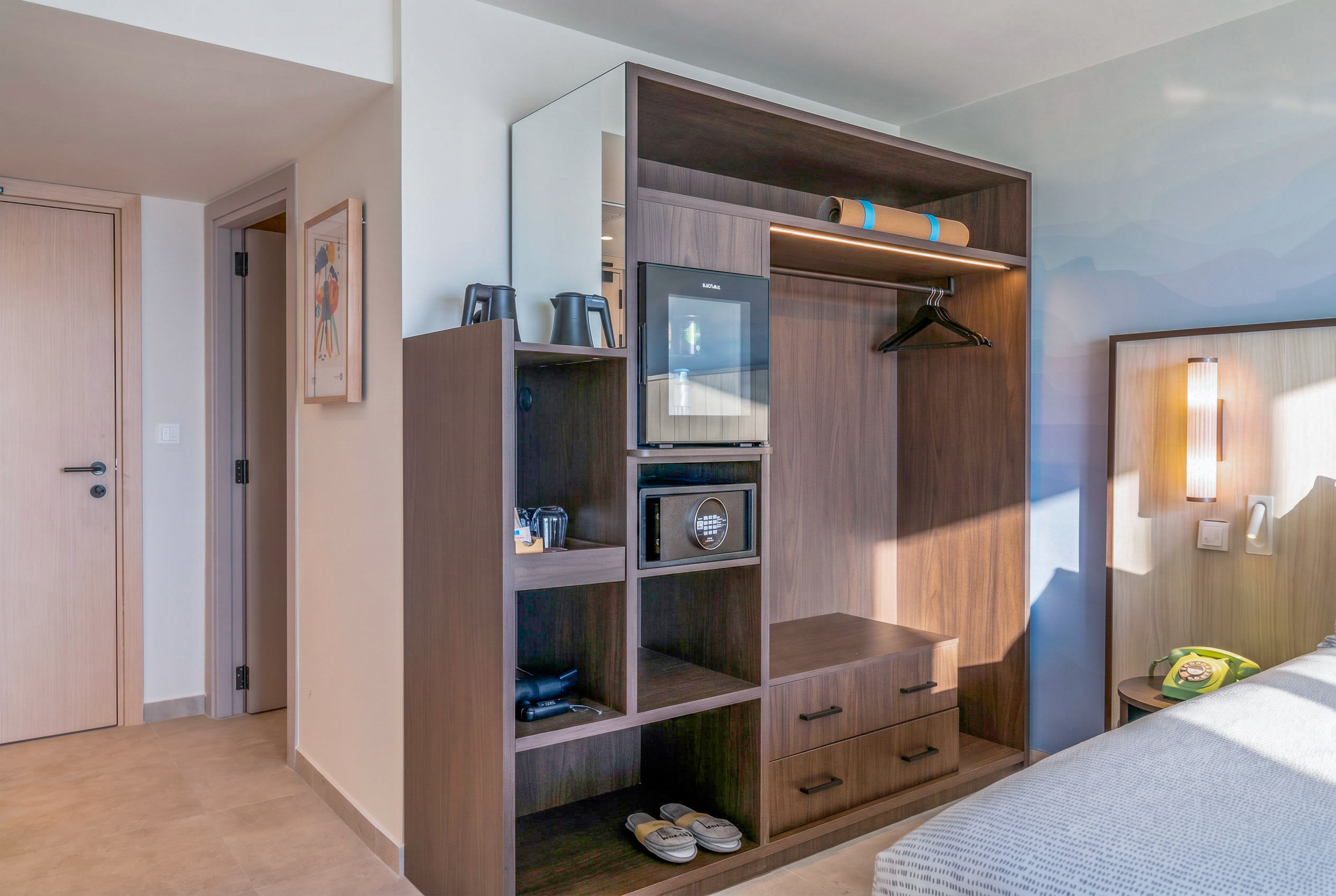 Modern hotel room with built-in dark wood wardrobe, mini-fridge, and safe.