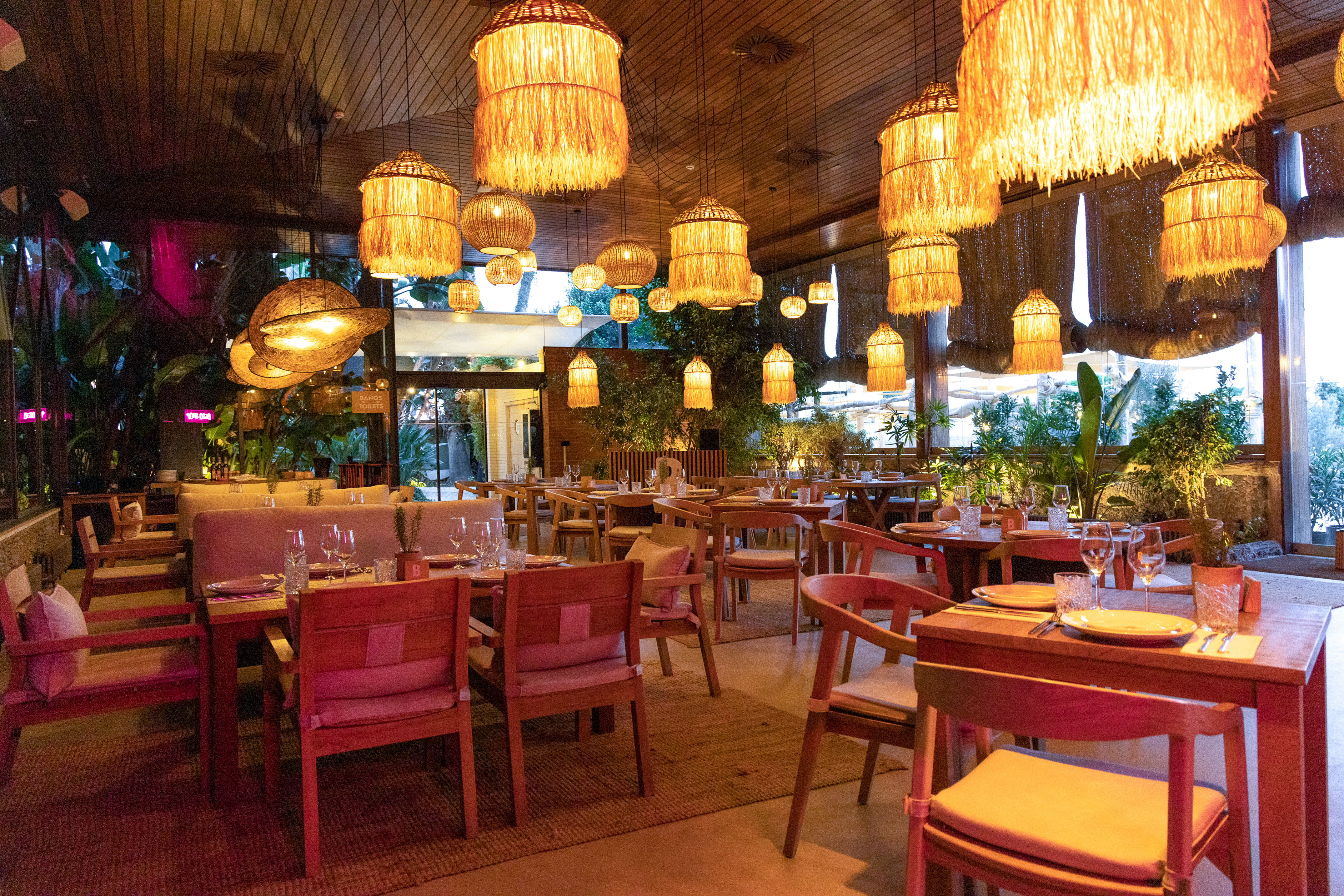 Warmly lit restaurant with hanging rattan lights, wooden tables & plants.