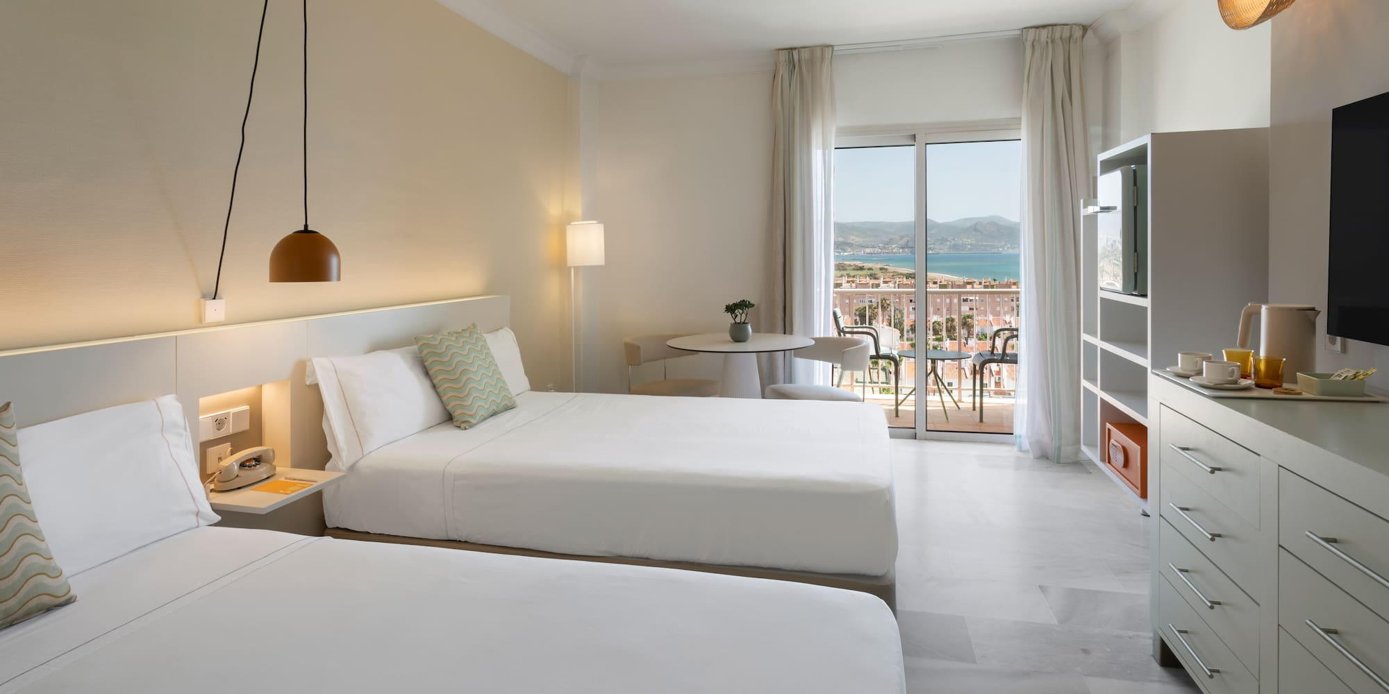 Amplia habitación de hotel con dos camas, balcón con vista al mar y montaña.