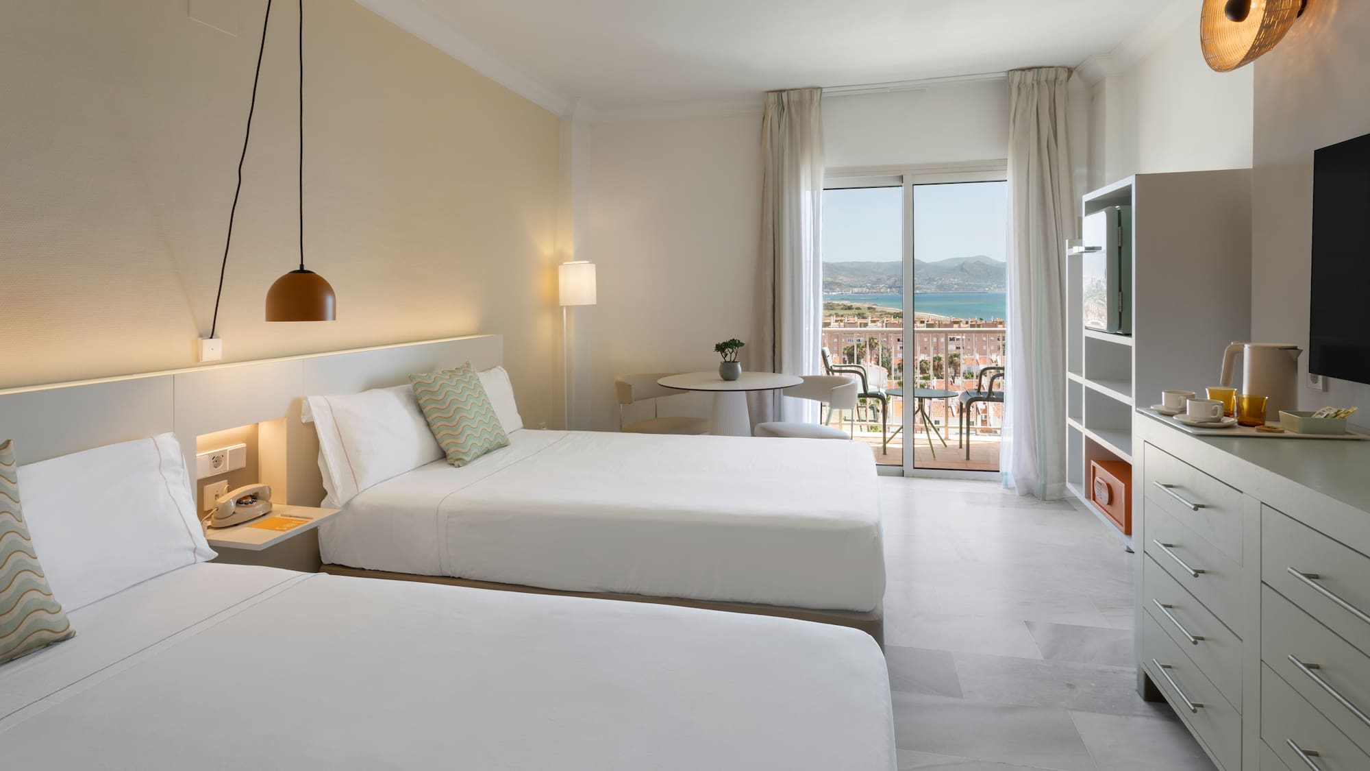 Amplia habitación de hotel con dos camas, balcón con vista al mar y montaña.