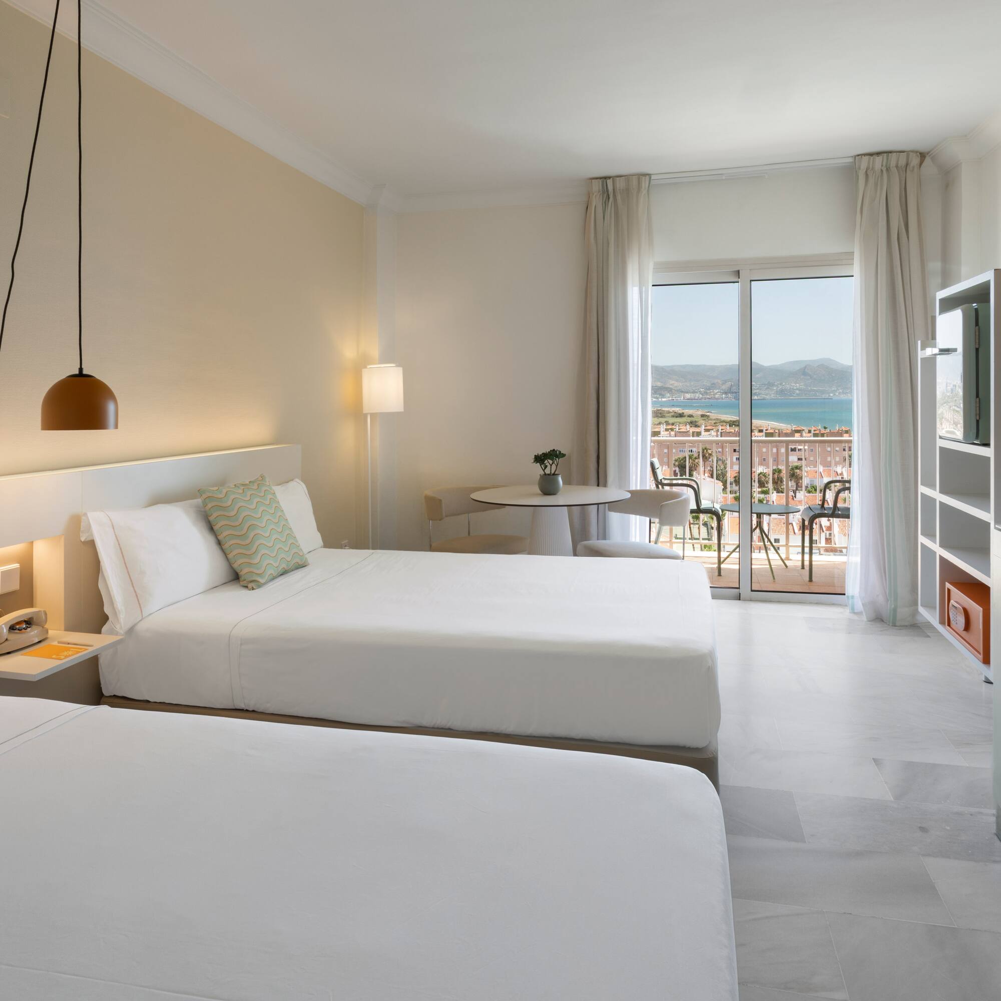 Amplia habitación de hotel con dos camas, balcón con vista al mar y montaña.