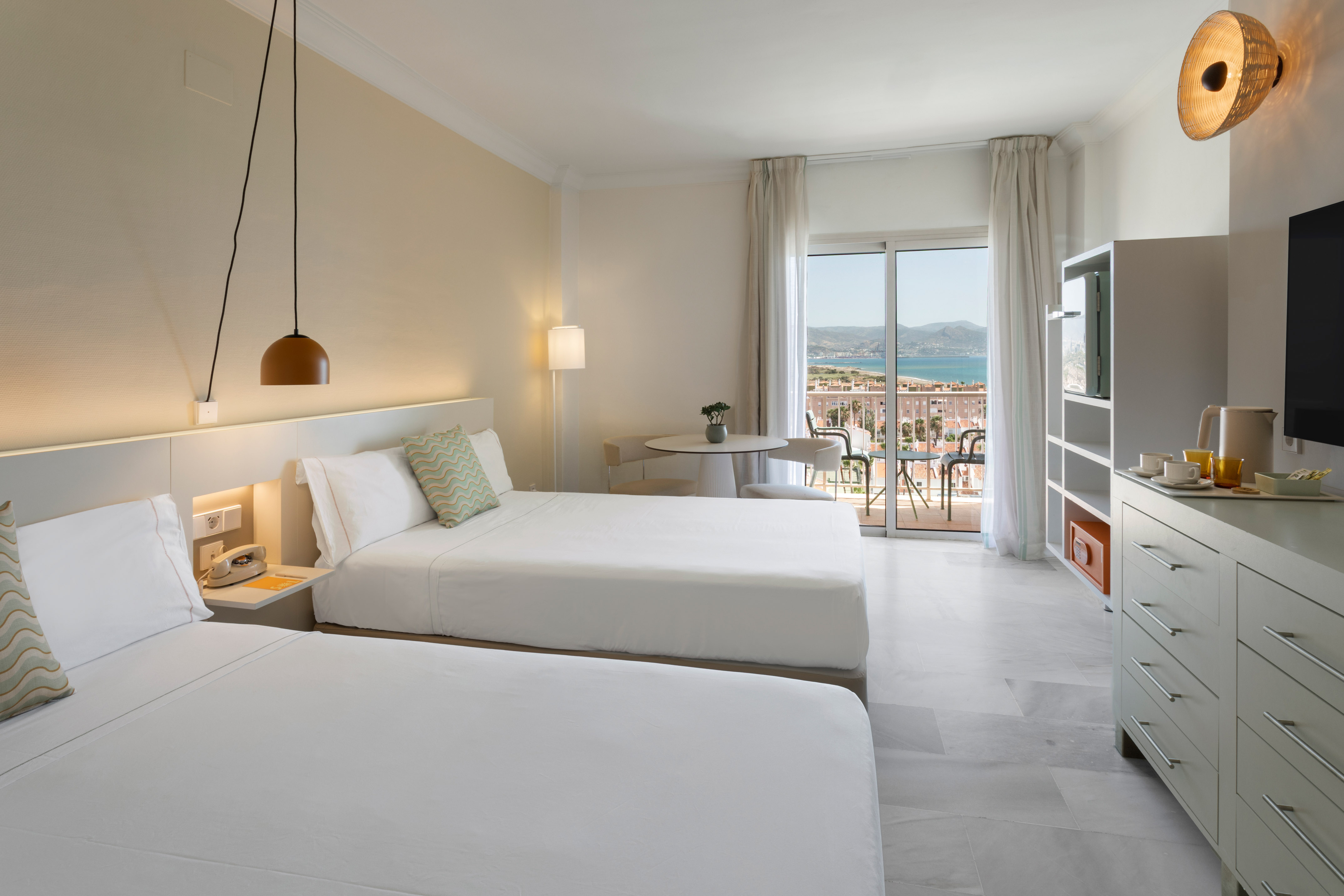 Amplia habitación de hotel con dos camas, balcón con vista al mar y montaña.