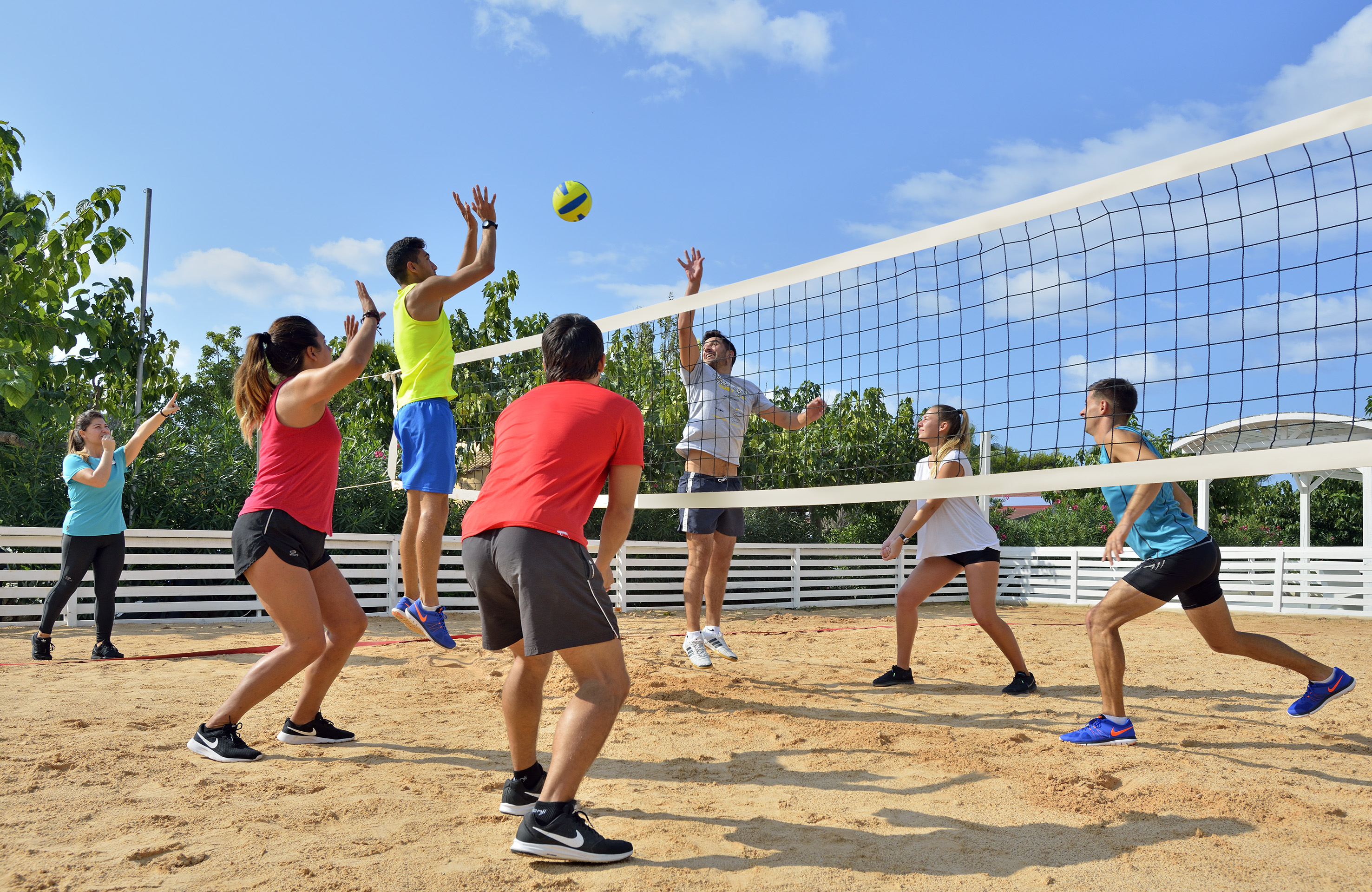 Six people playing beach volleyball on a sunny sand court with a net.