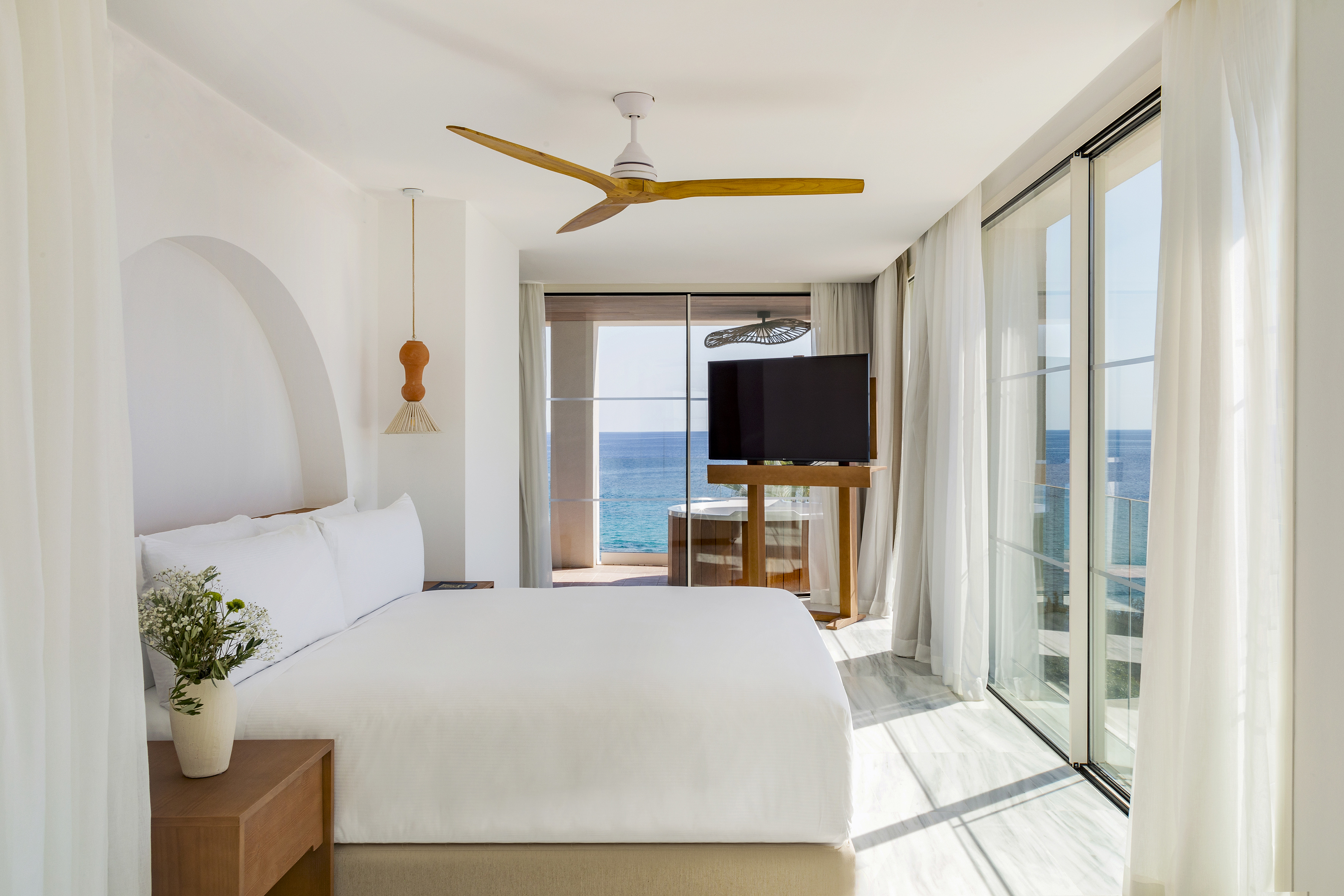 Resort bedroom with ocean view balcony, white bed, and modern wooden fan.