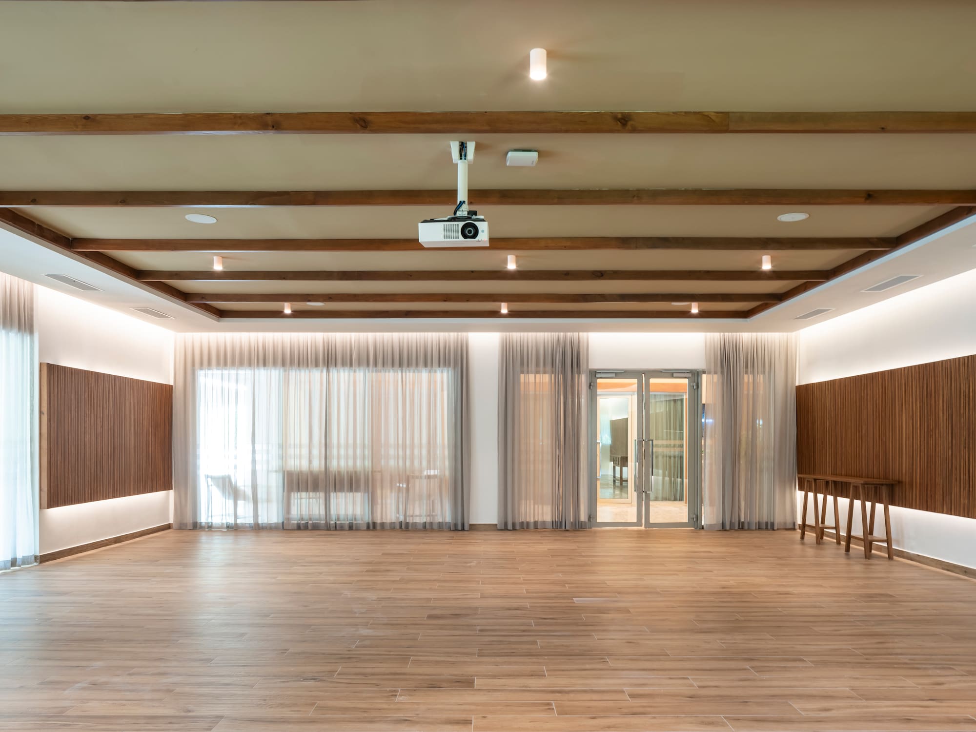 Modern event room with wood flooring, ceiling projector, and sheer curtains.