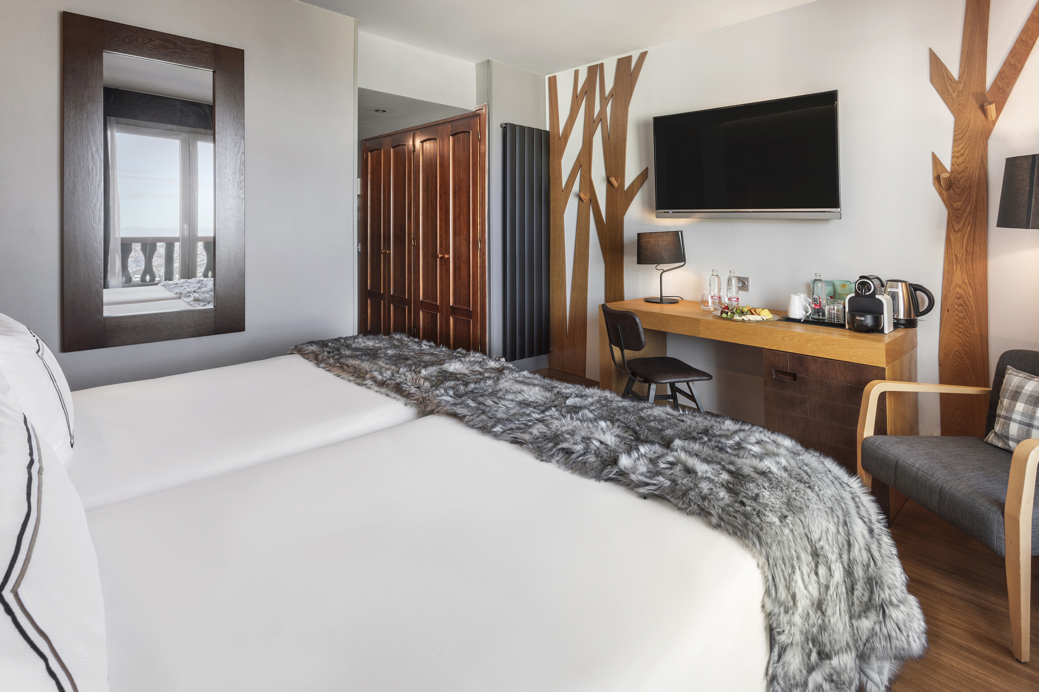 Modern hotel room with twin beds, desk, TV, and tree-inspired wall art.