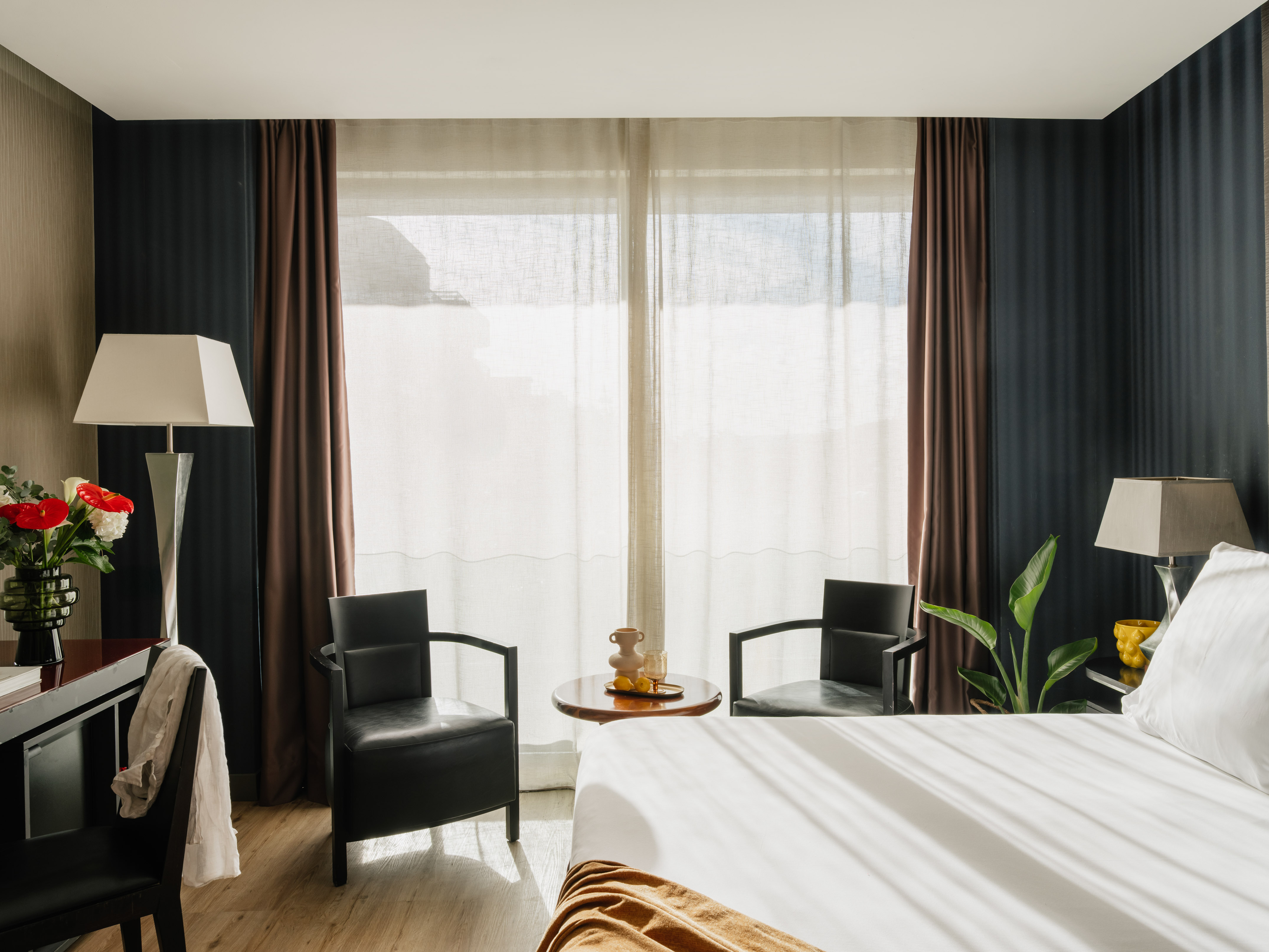 Modern hotel bedroom with white bed, seating, desk, and large window.