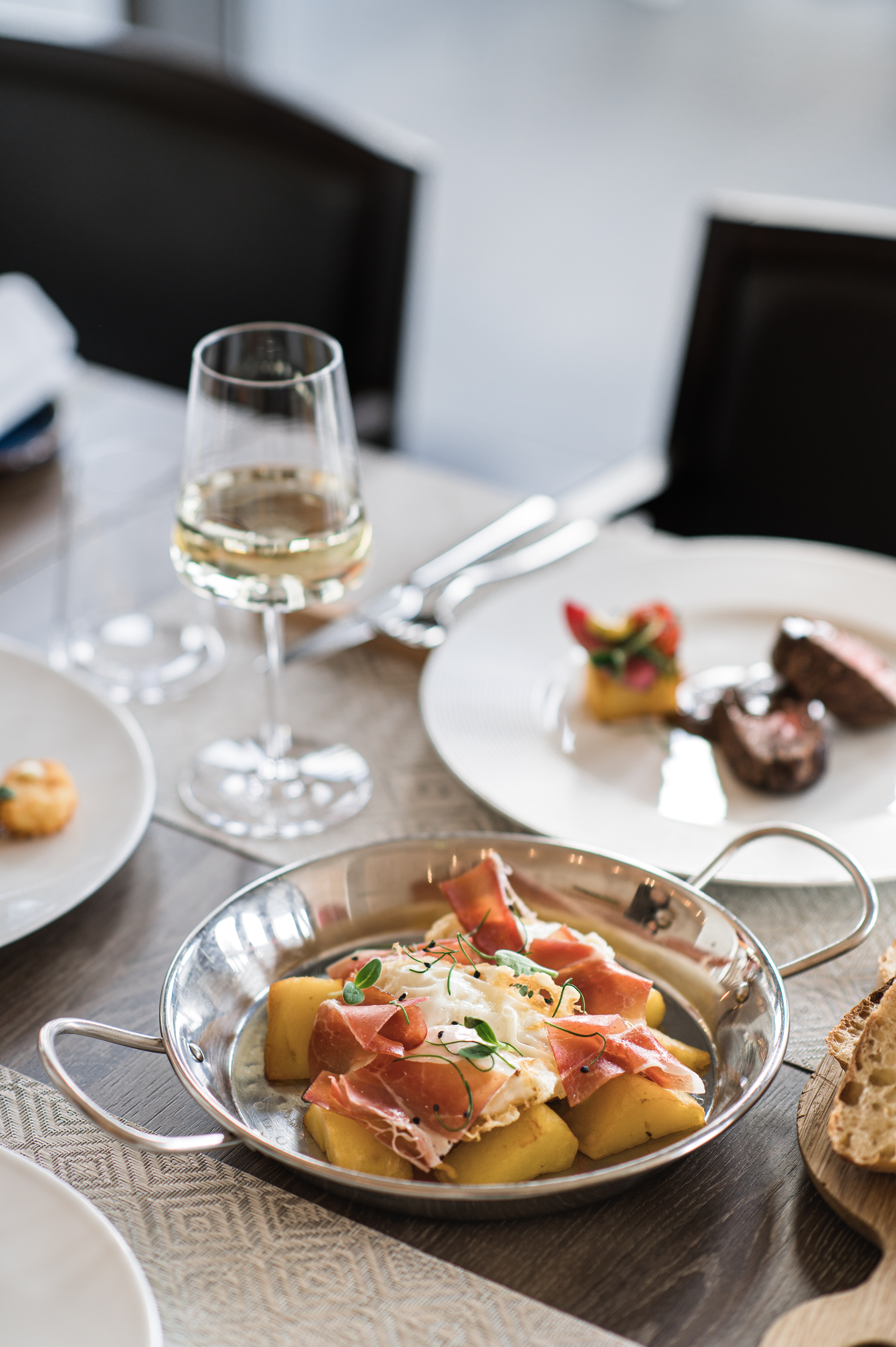 Skillet of fried eggs, prosciutto, potatoes; white wine on a restaurant table.
