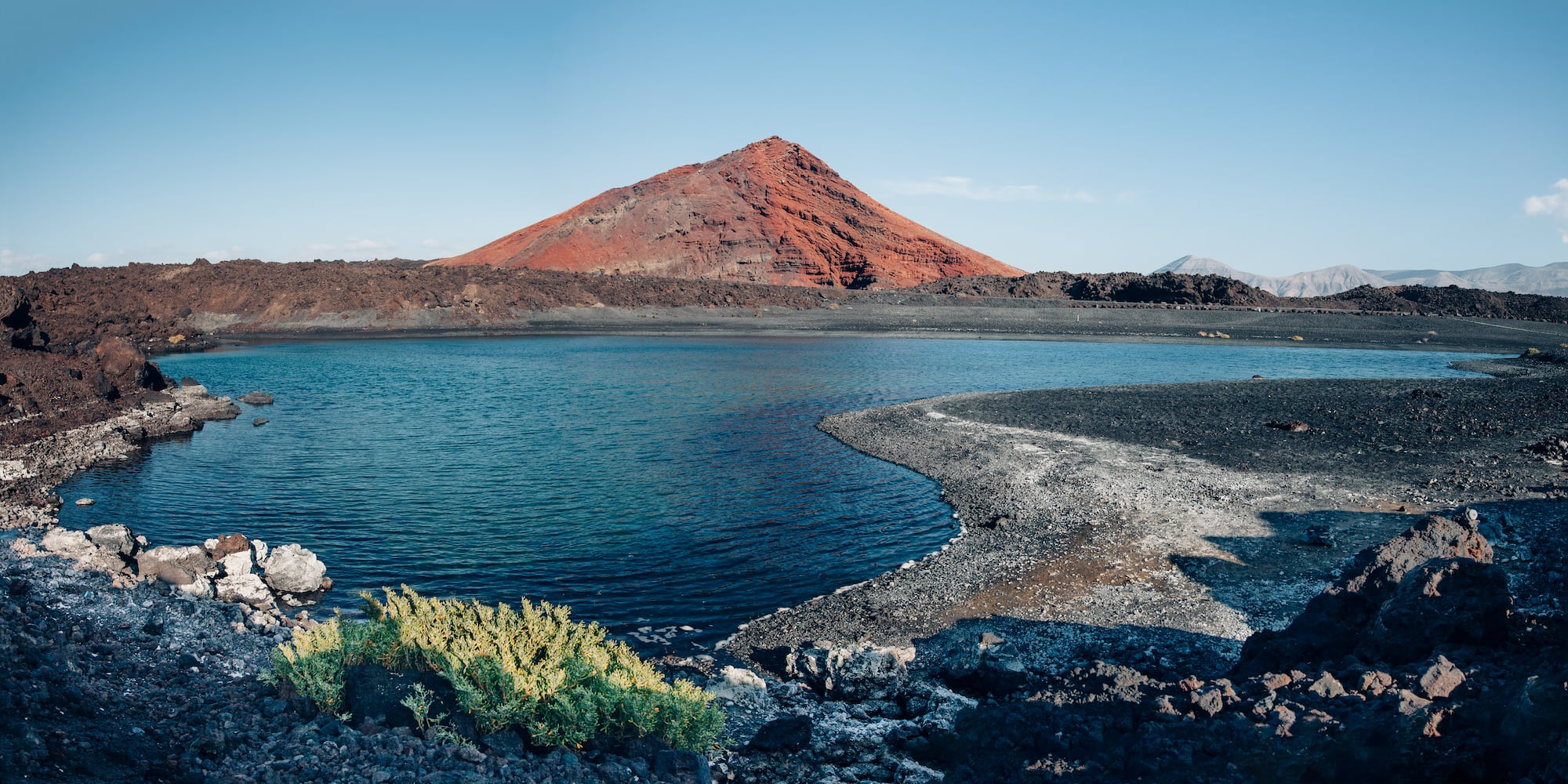 Red volcanic mountain overlooking a blue lake with black sand shore.