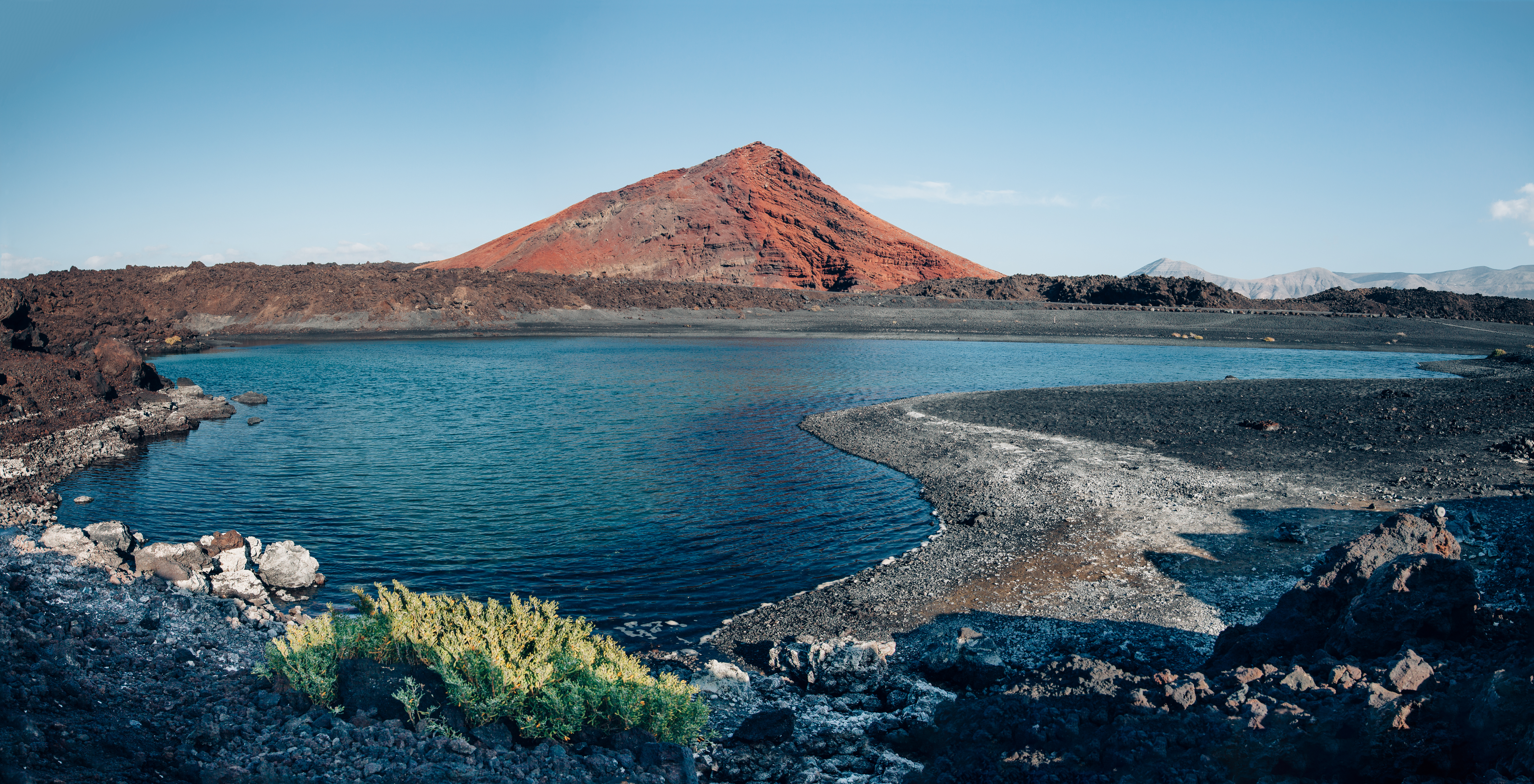 Red volcanic mountain overlooking a blue lake with black sand shore.