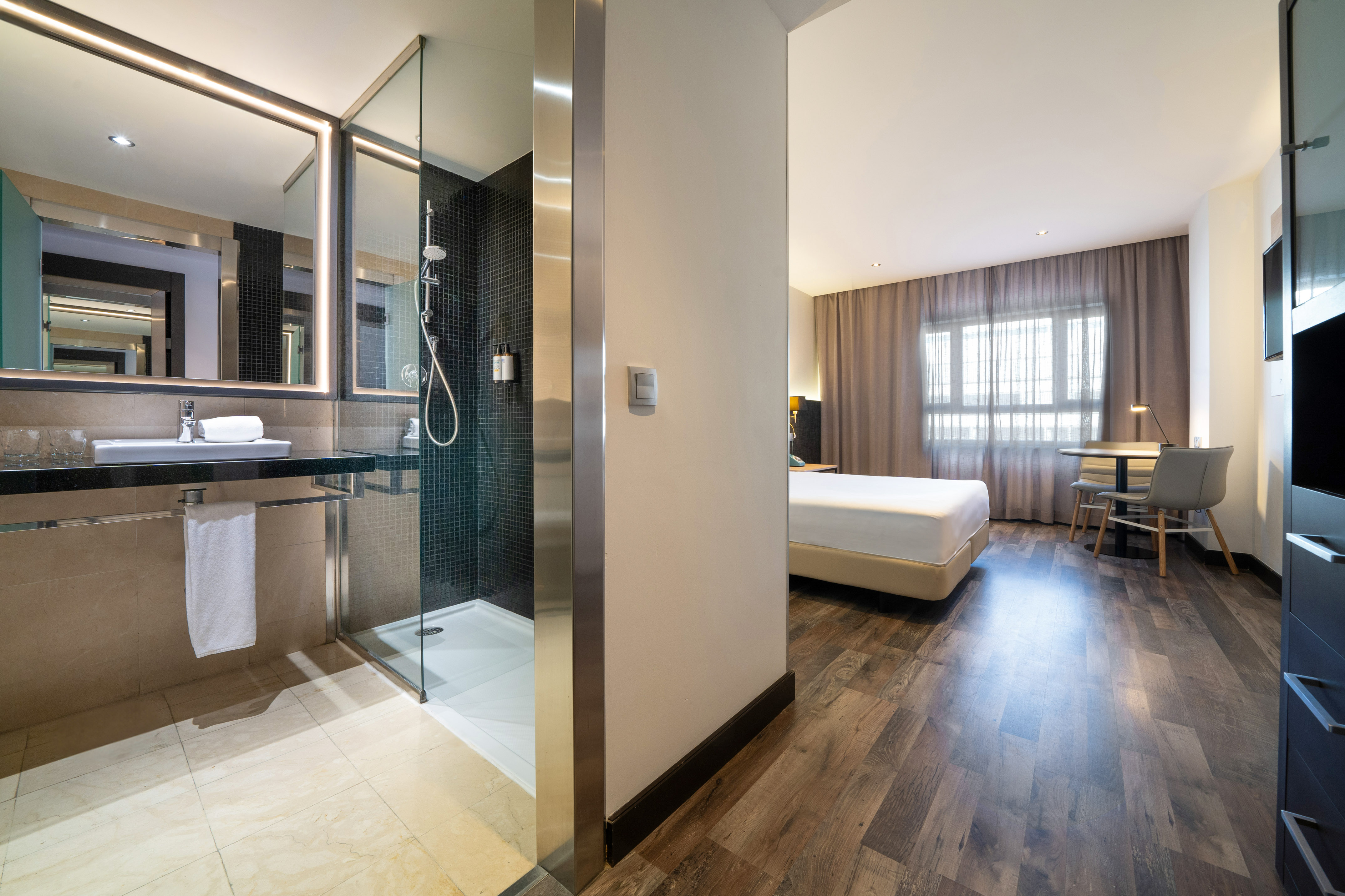 Modern hotel room with open bathroom, glass shower, bed, and desk.
