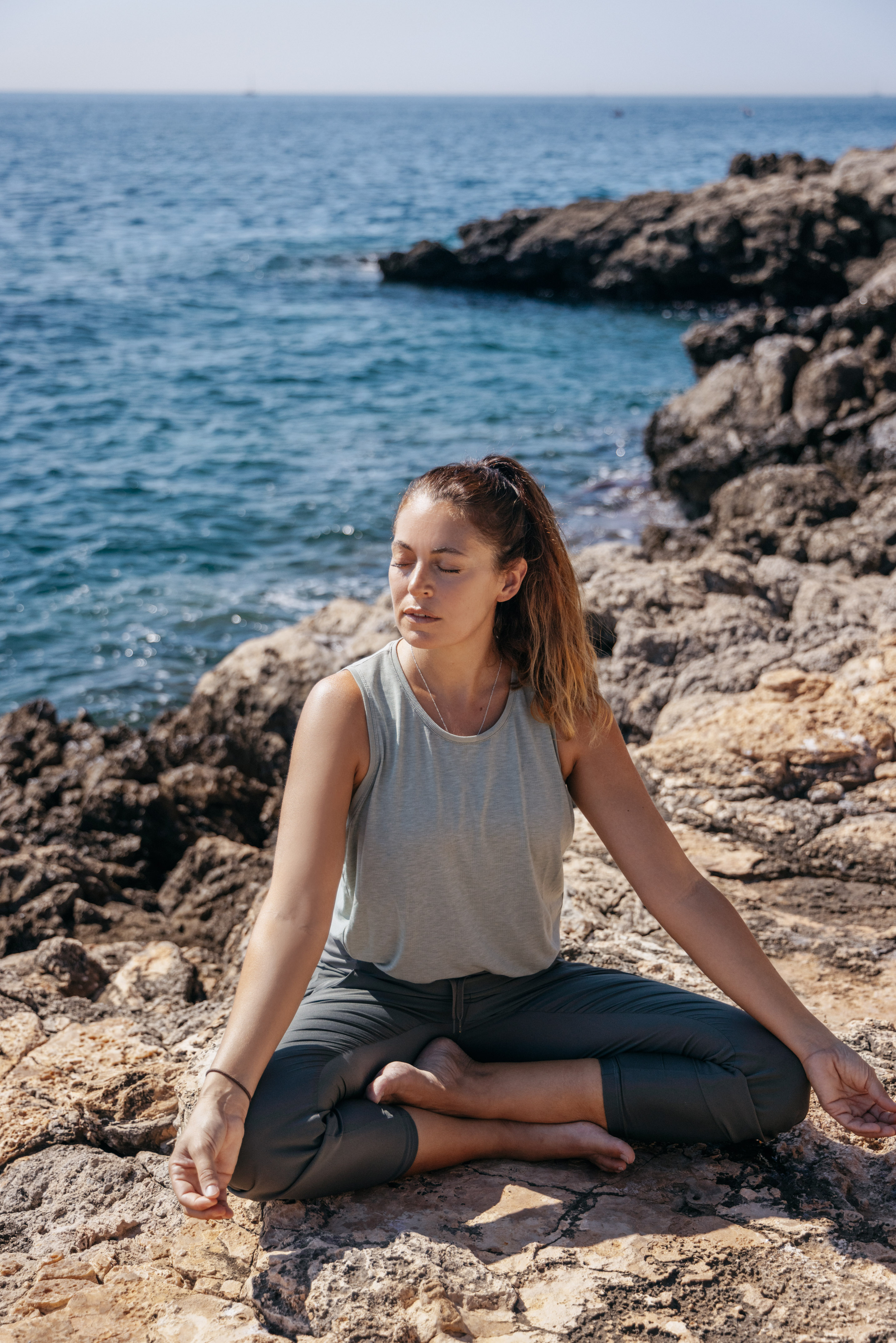 Woman in yoga meditation pose on sunny rocky coast by the ocean.