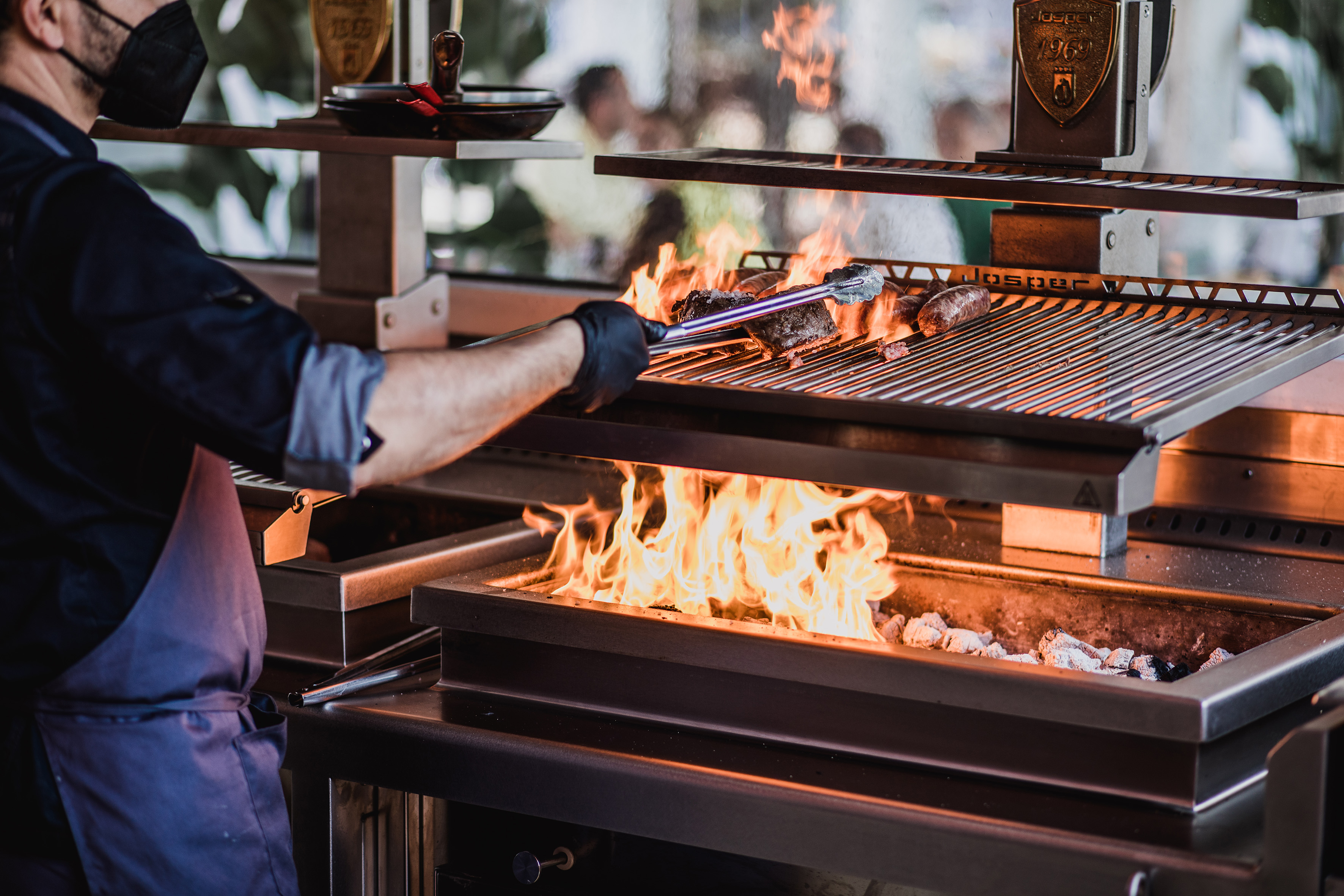 Chef in mask grilling steak on a Josper barbecue with roaring flames.