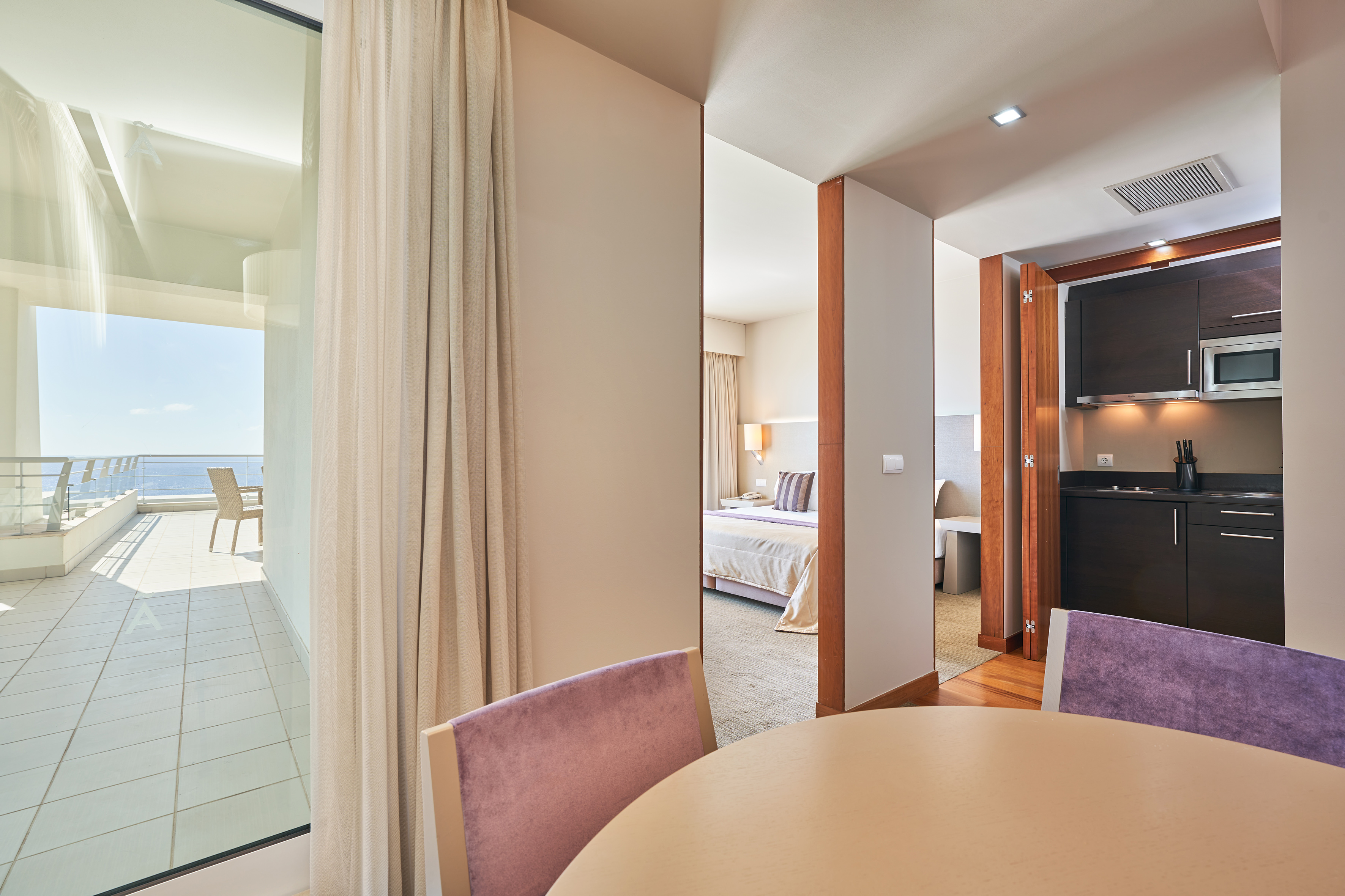 Hotel suite with ocean view balcony, dining area, bedroom, and kitchenette.