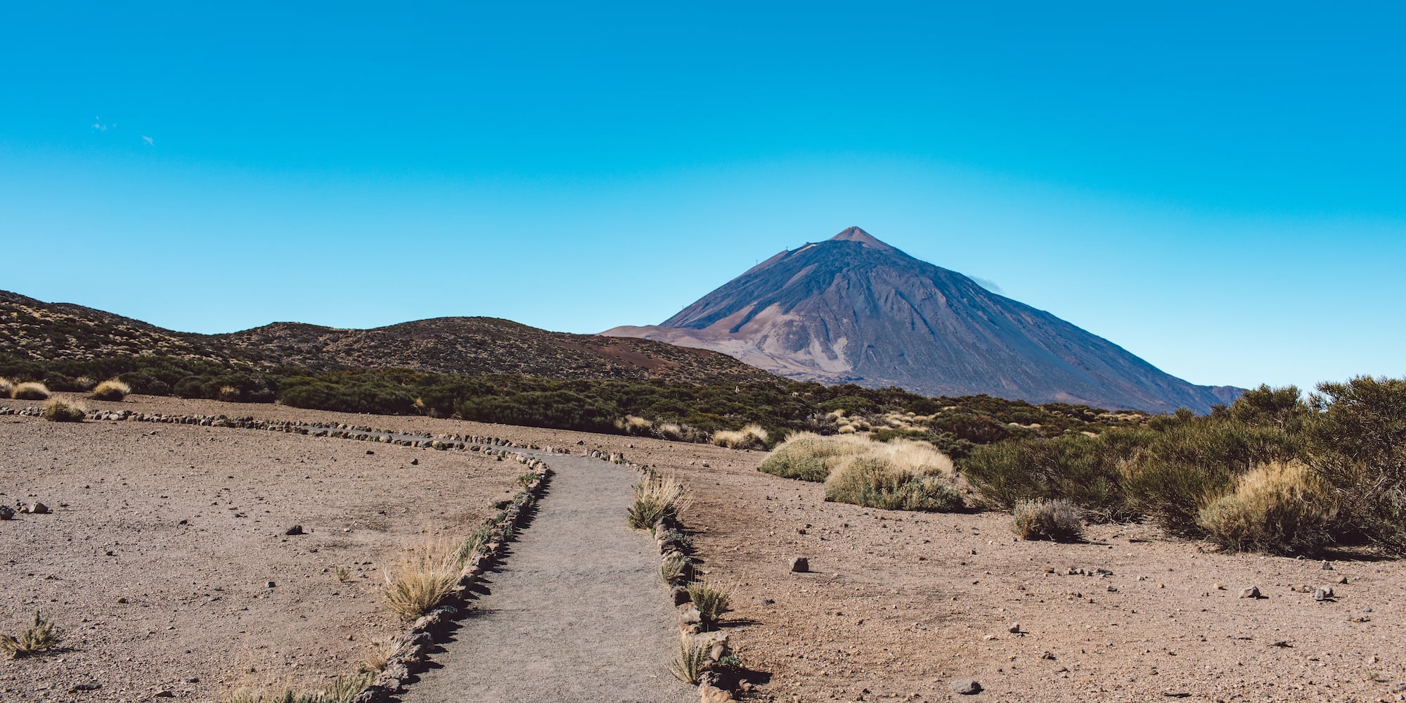 Gravel path to Teide volcano, clear blue sky over Tenerife volcanic landscape.