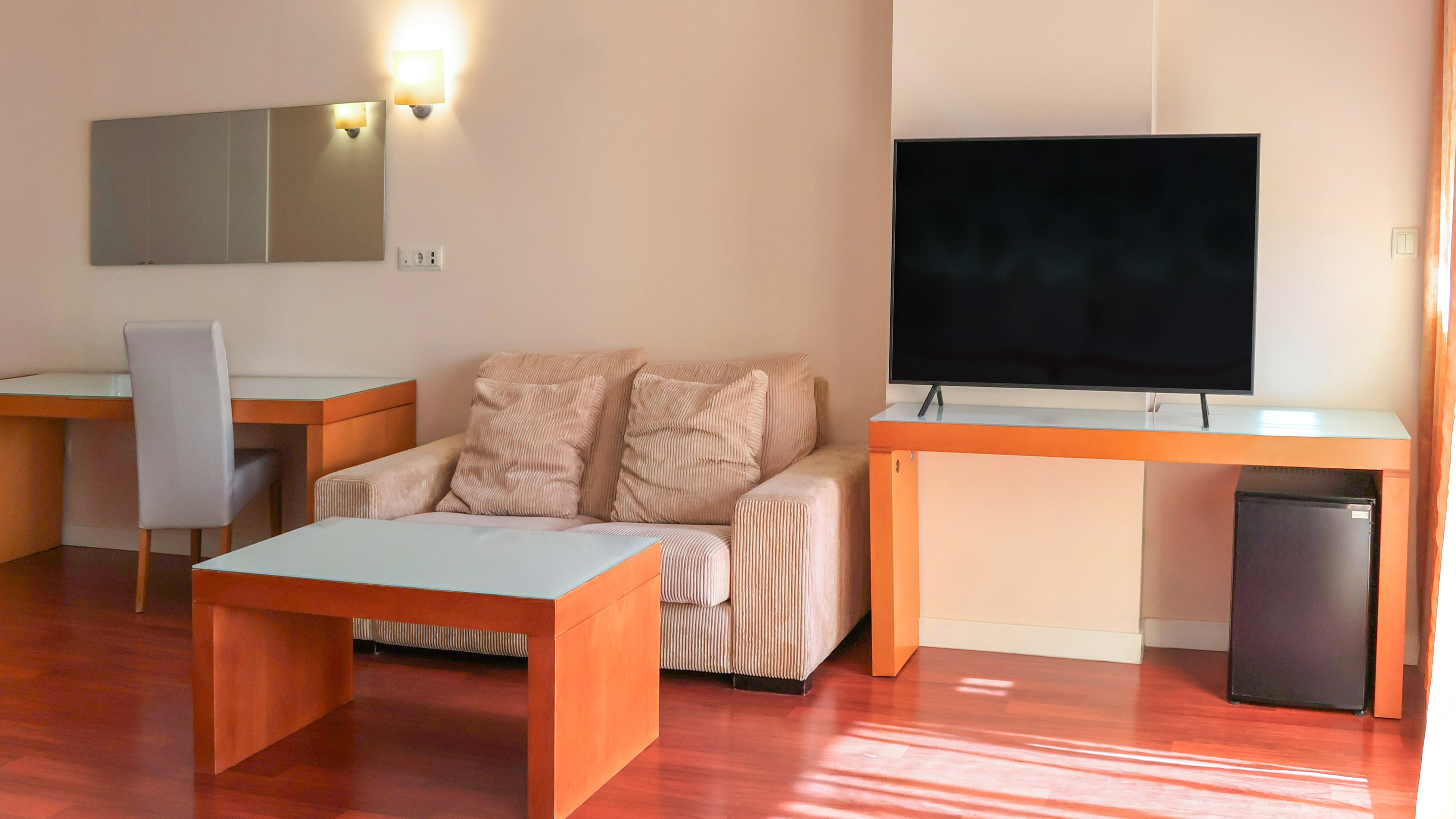 Modern hotel room living area with sofa, flat-screen TV, desk, and wood floor.