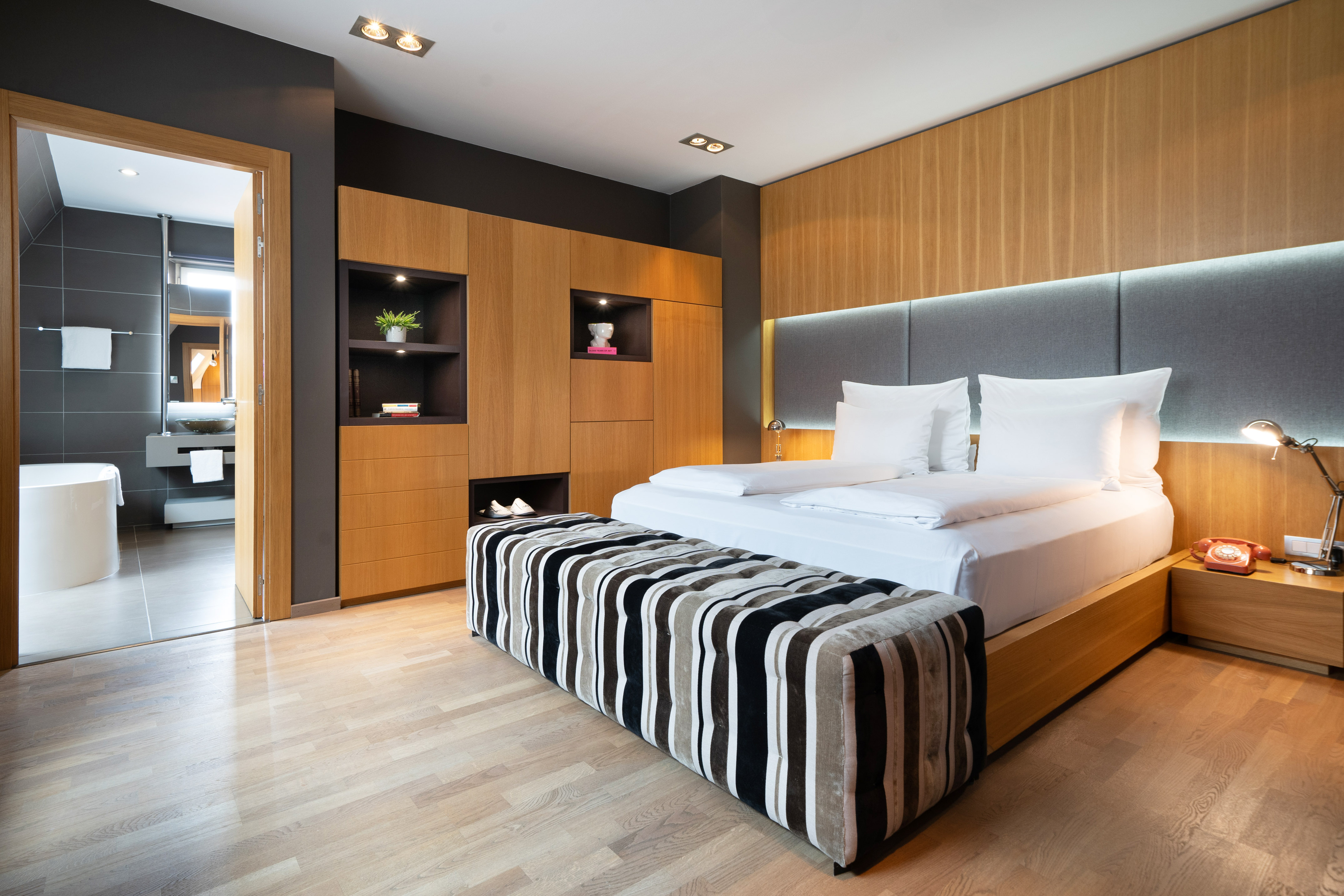 Modern hotel bedroom with wooden panels, king bed, striped ottoman, and ensuite bath.