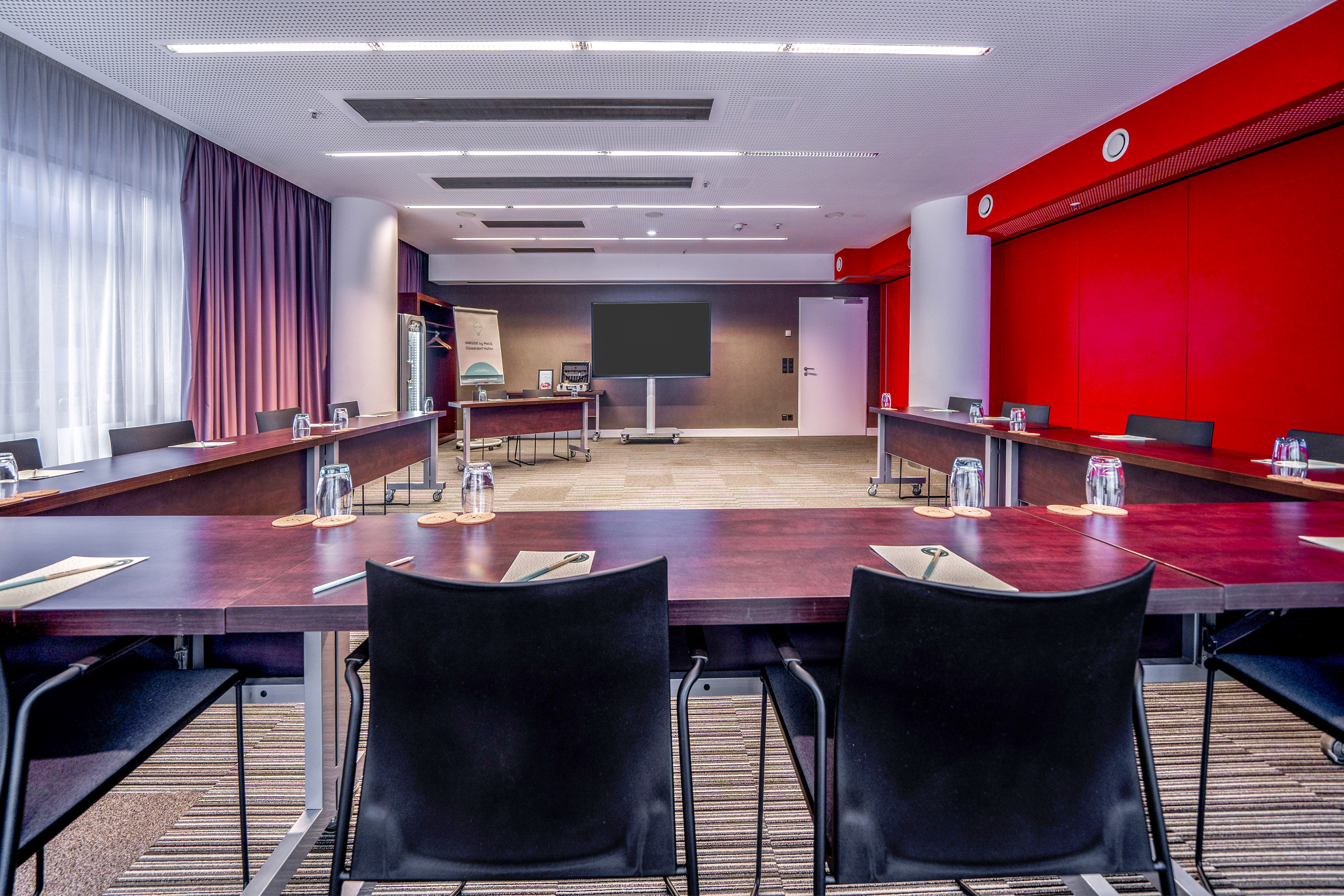 Modern hotel meeting room with U-shape table, large TV, and red wall accents.