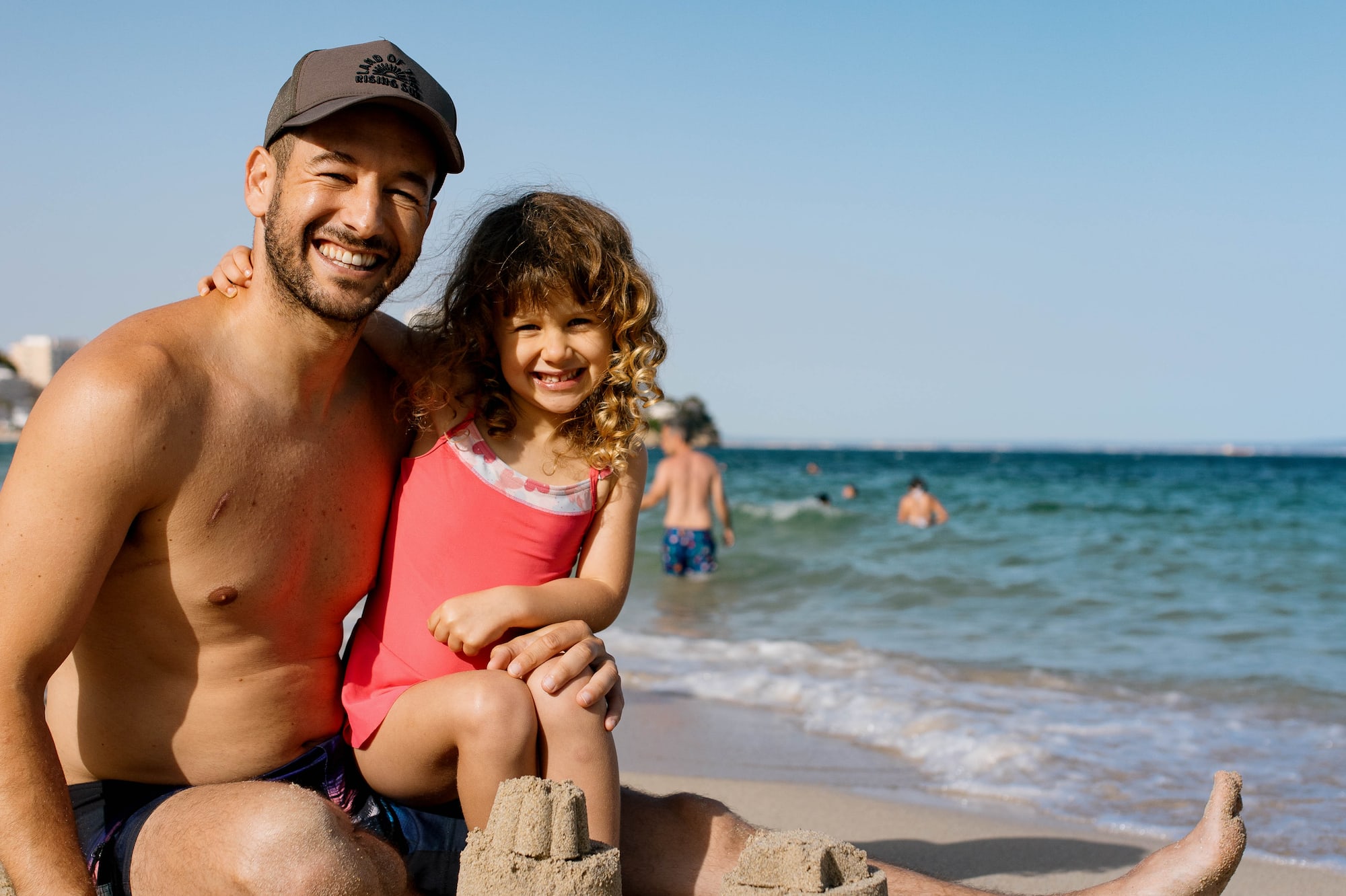 Padre e hija sonrientes en la playa con castillos de arena y mar.