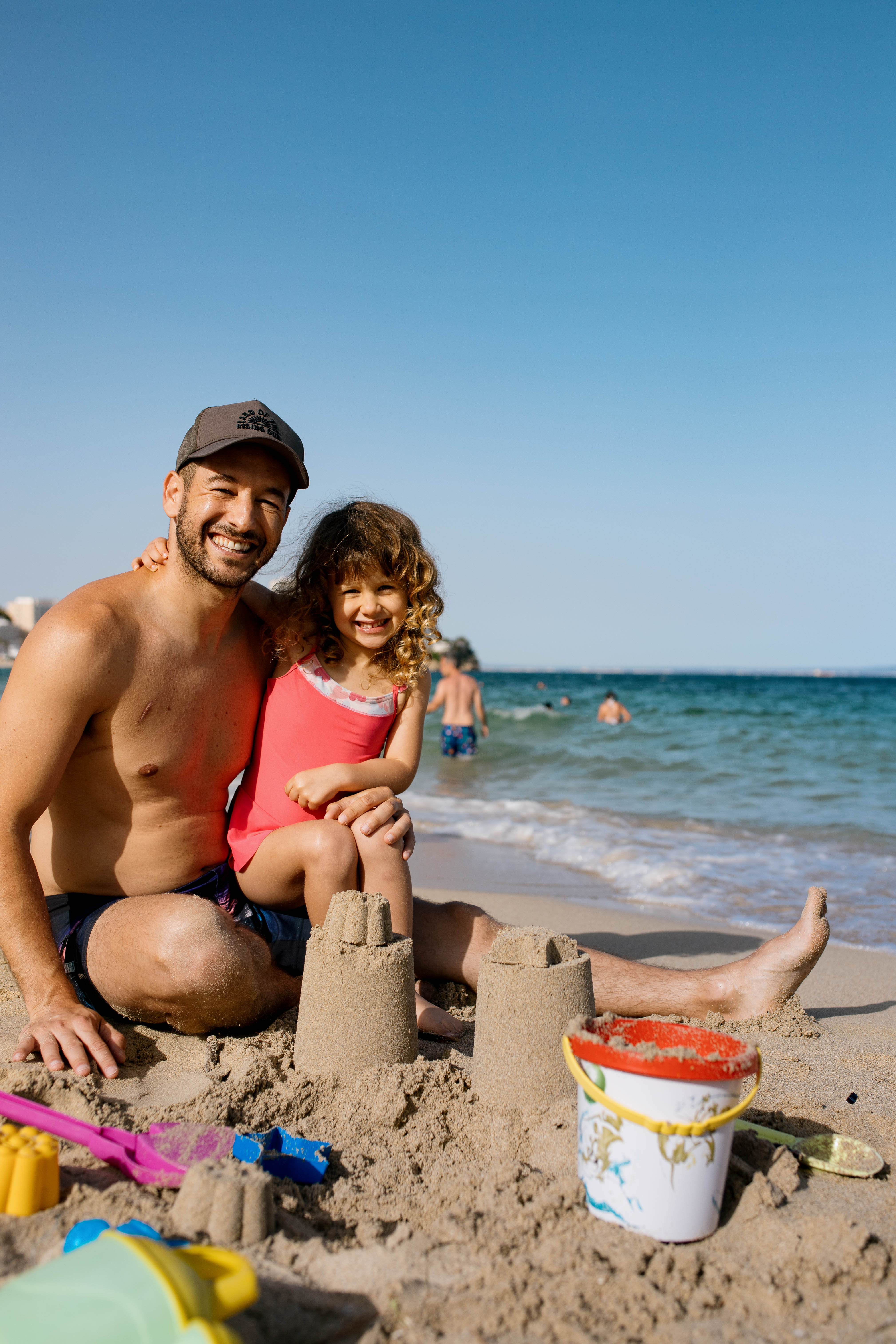 Padre e hija sonrientes en la playa con castillos de arena y mar.